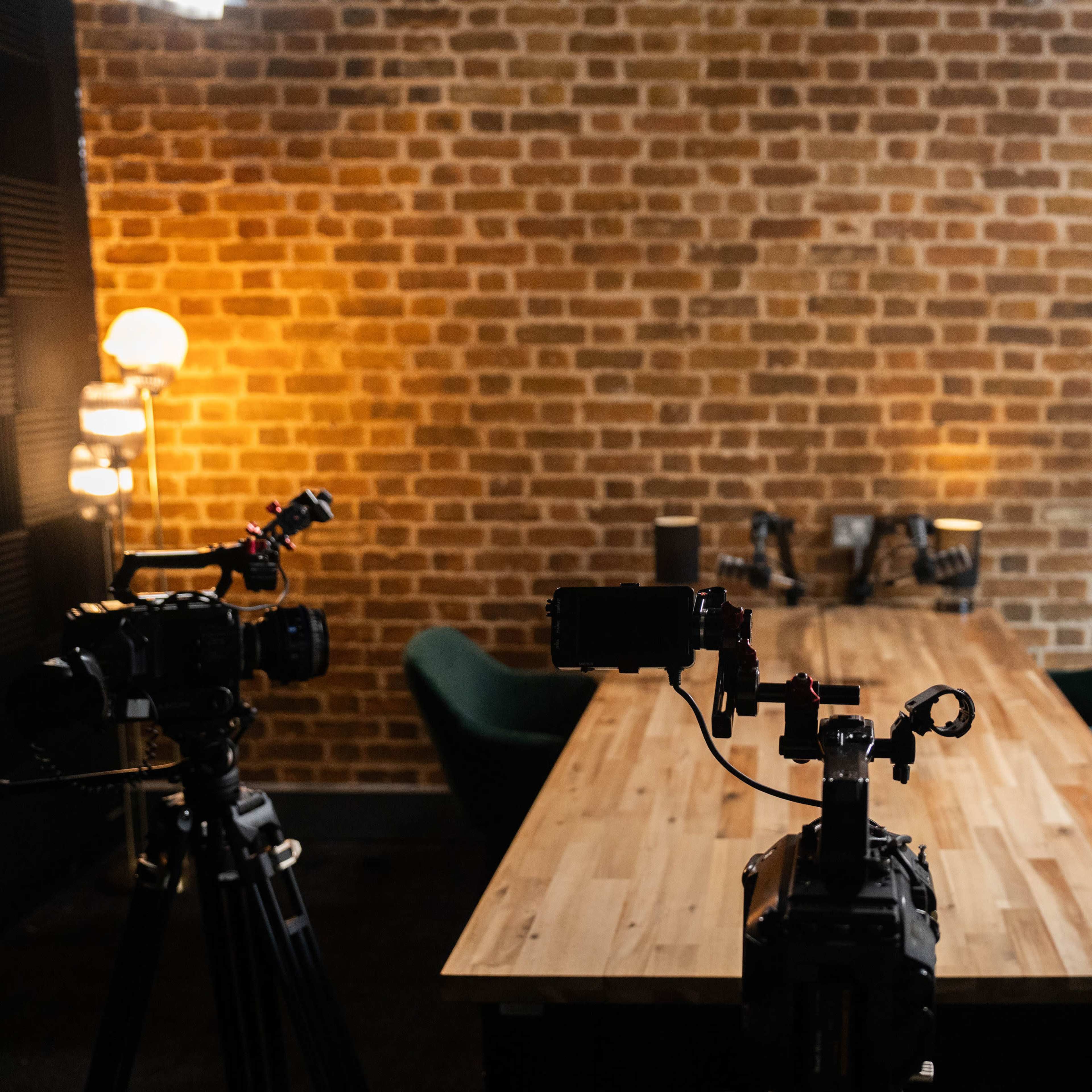 A well-lit, spacious room with a wooden table and three cameras positioned on tripods facing the table, against a brick wall backdrop.