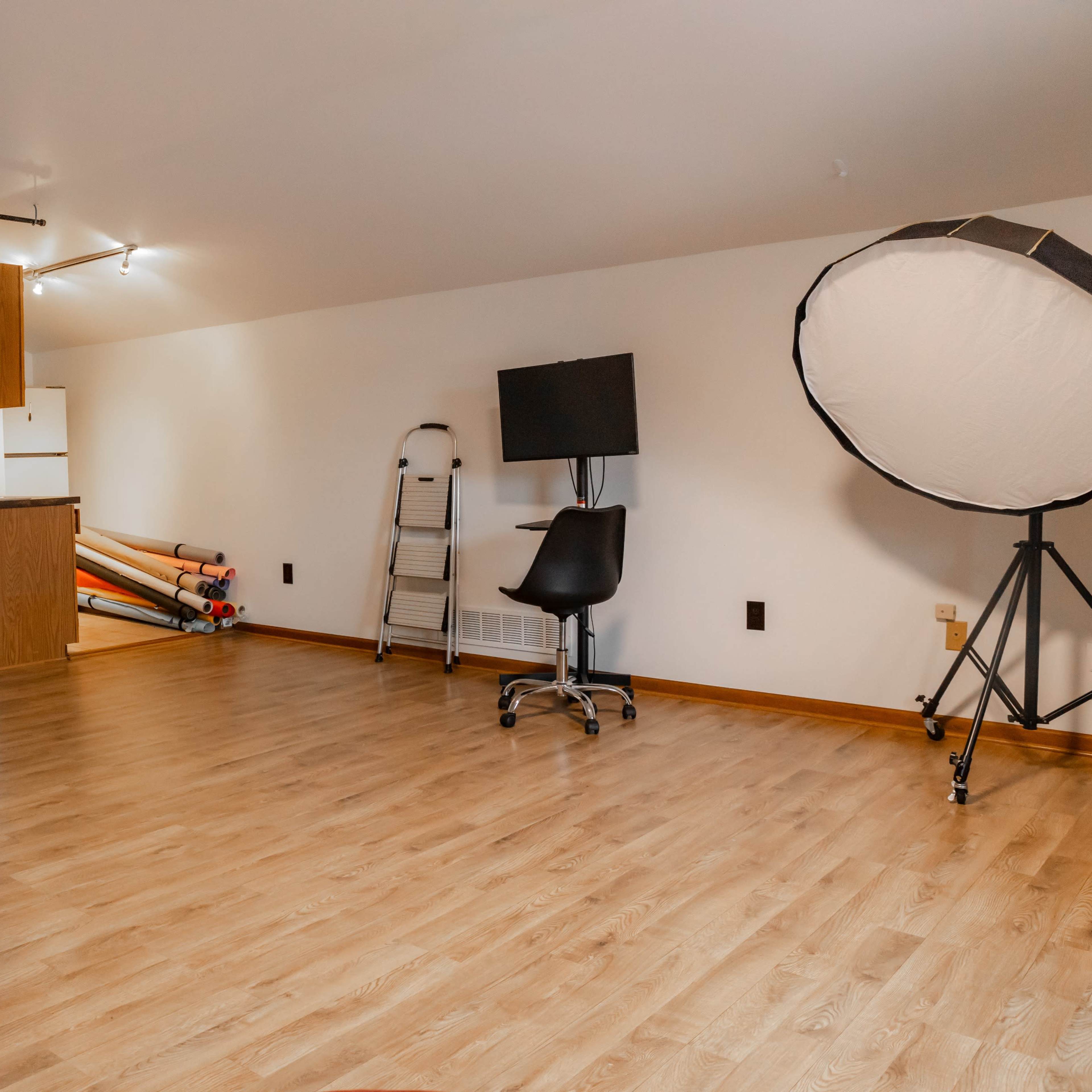 An empty room with wooden flooring, a kitchenette in the background, a tall standing light, a black chair on wheels, a folding ladder, and rolls of backdrop paper against the wall.