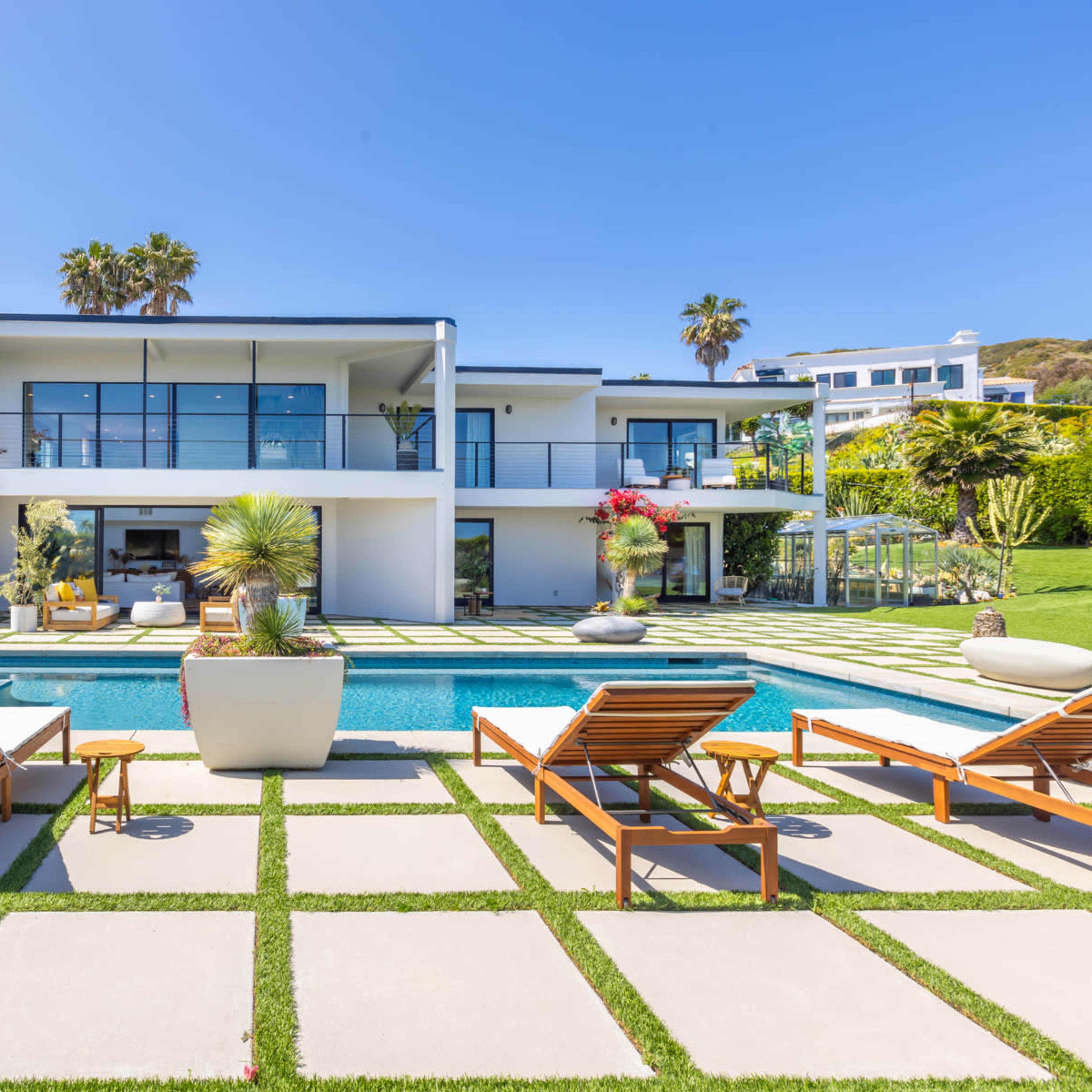 A modern two-story house with a swimming pool and lounge chairs is surrounded by landscaped gardens and palm trees.