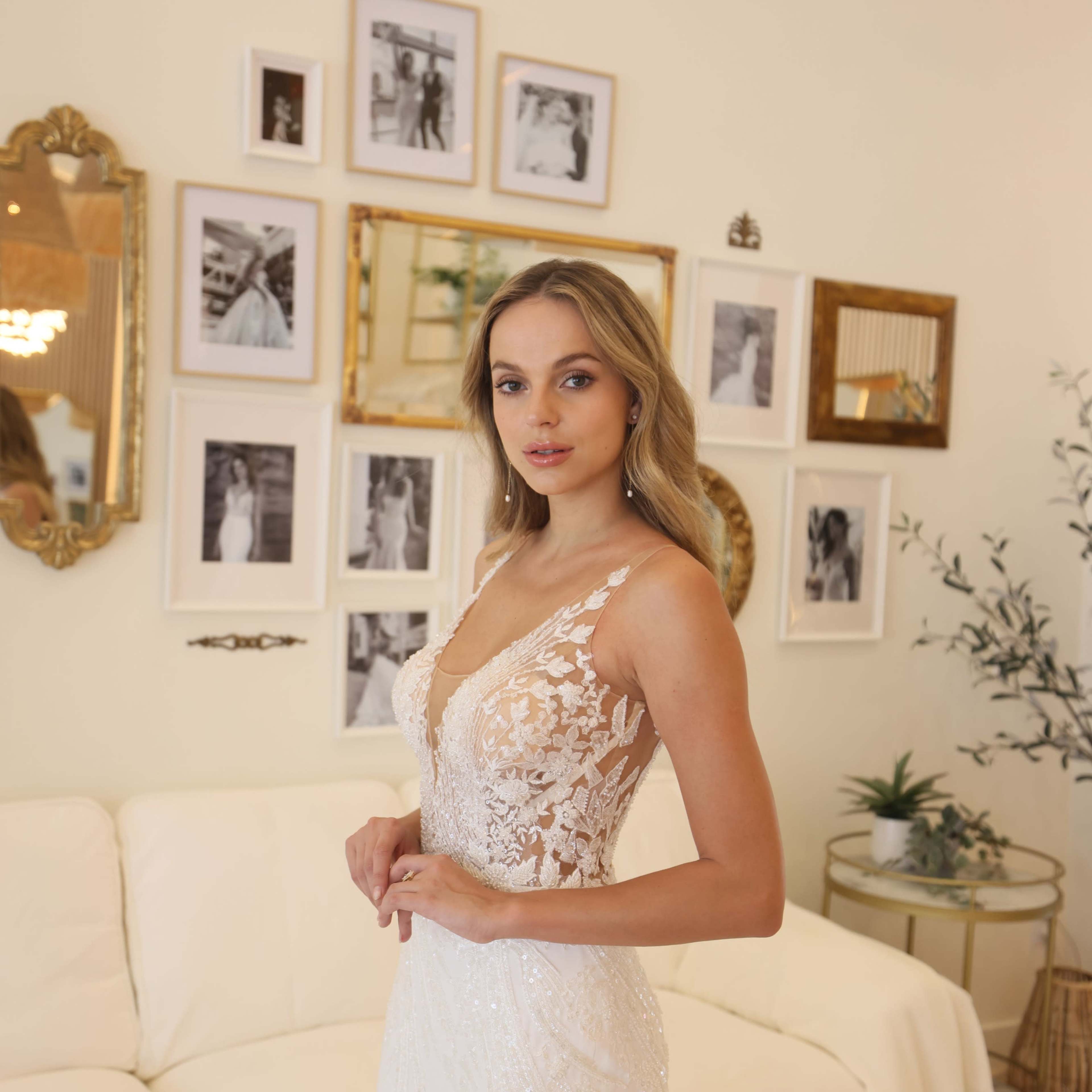 A woman in a white bridal gown stands in a room decorated with framed black-and-white photos on the wall.