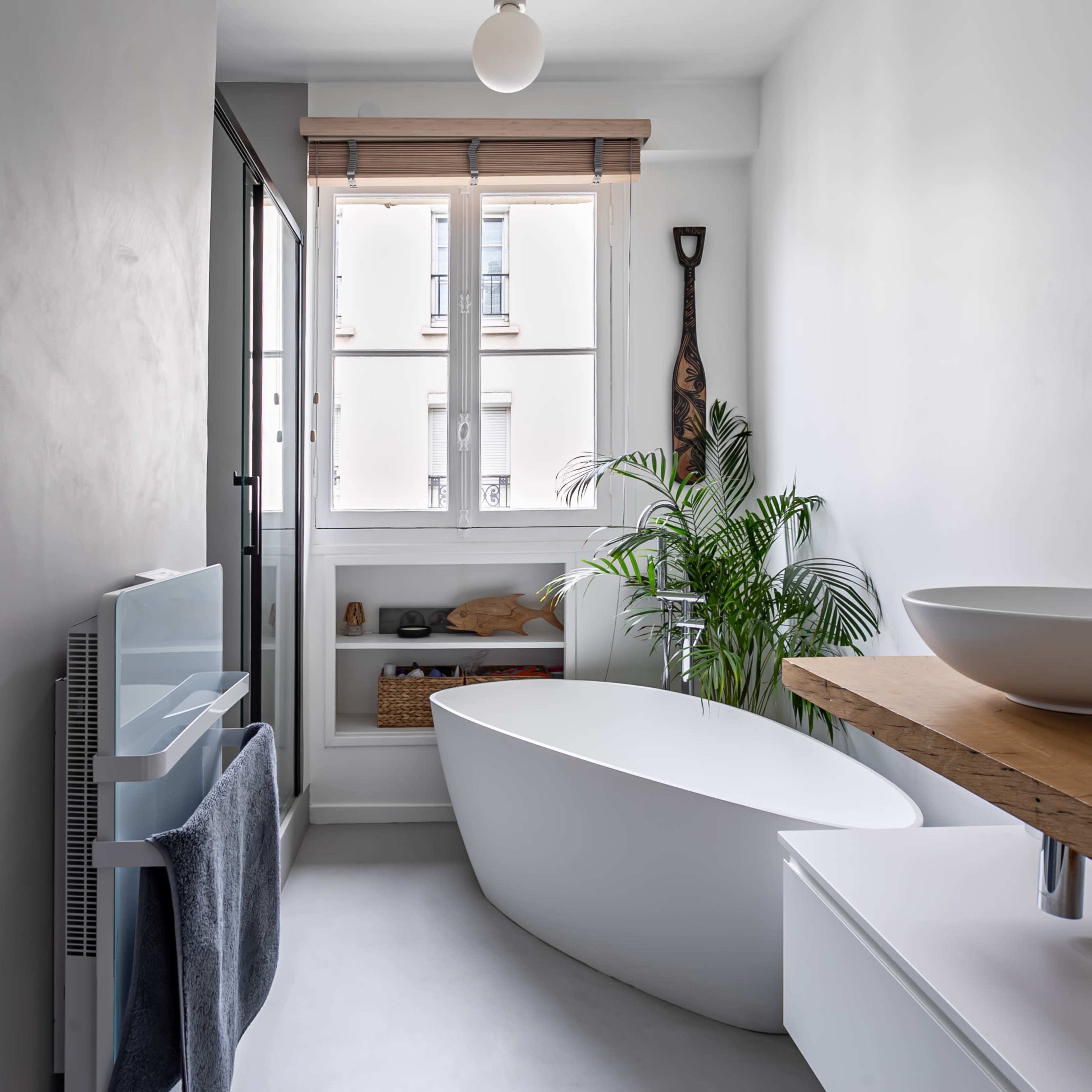 A modern bathroom features a freestanding tub, a wooden counter with a bowl sink, and large windows allowing natural light, complemented by indoor plants.
