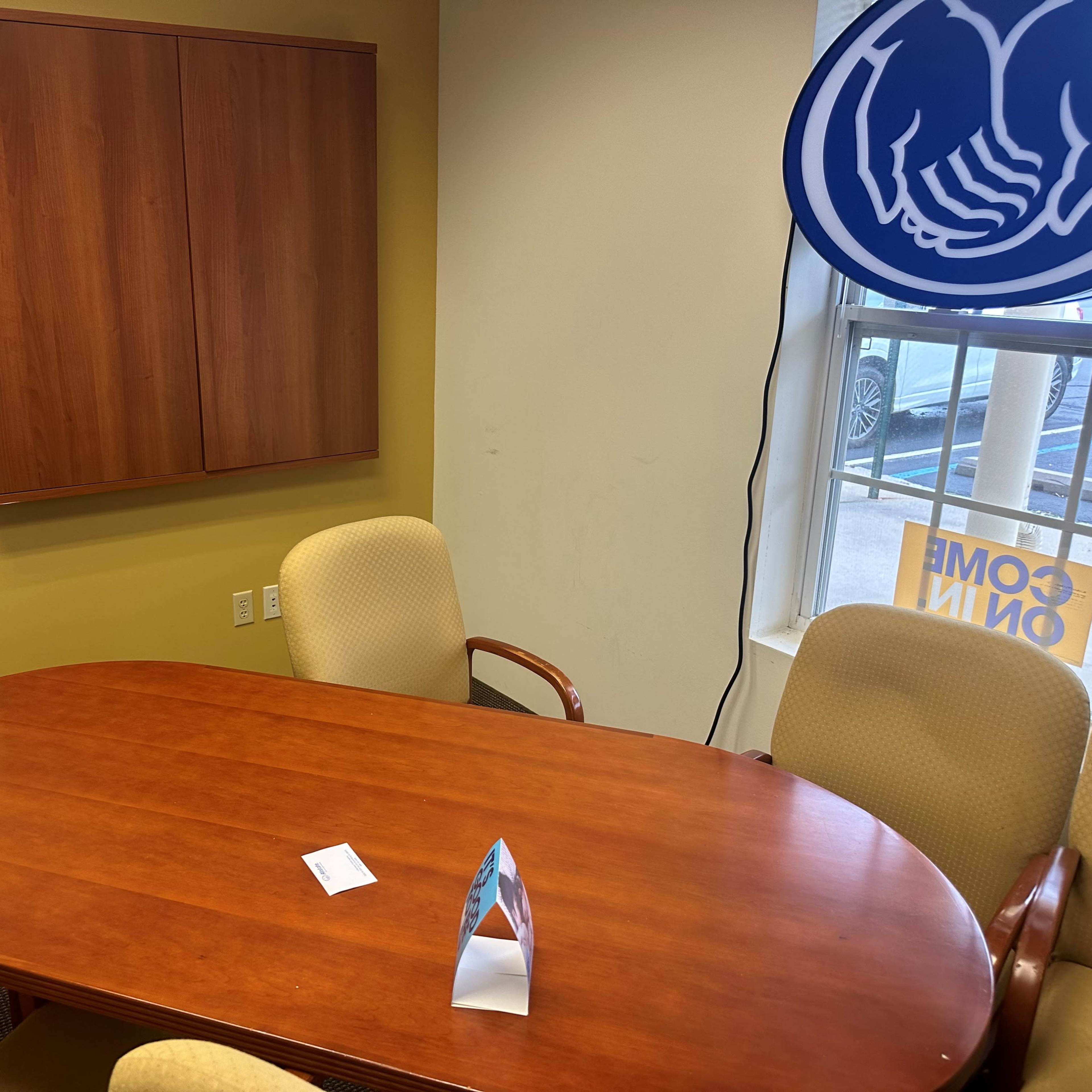 A wooden conference table surrounded by four chairs sits in a small office with a cabinet on the wall and a window displaying a sign.