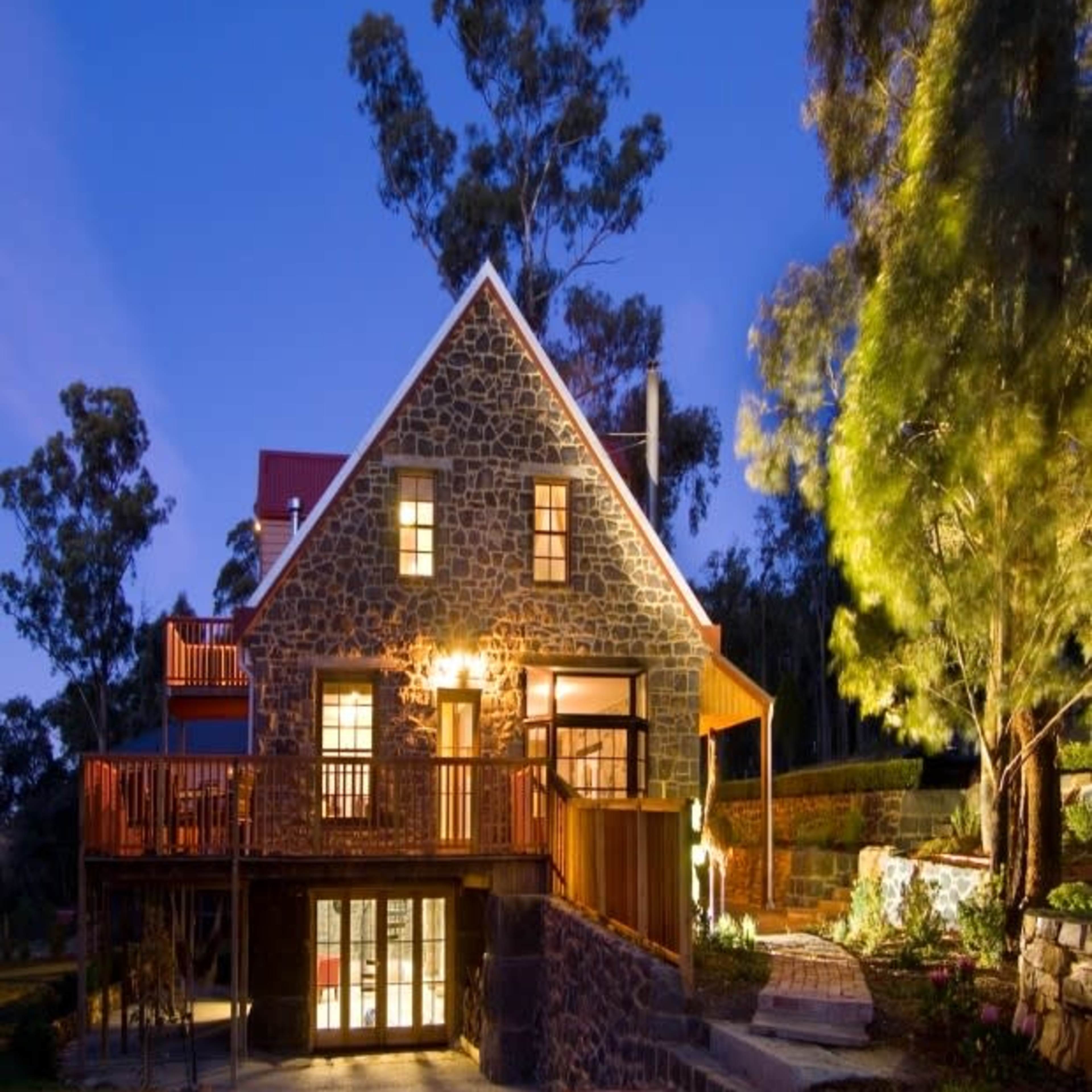 A stone house with a triangular roof and large windows is illuminated at dusk, surrounded by trees and outdoor landscaping.