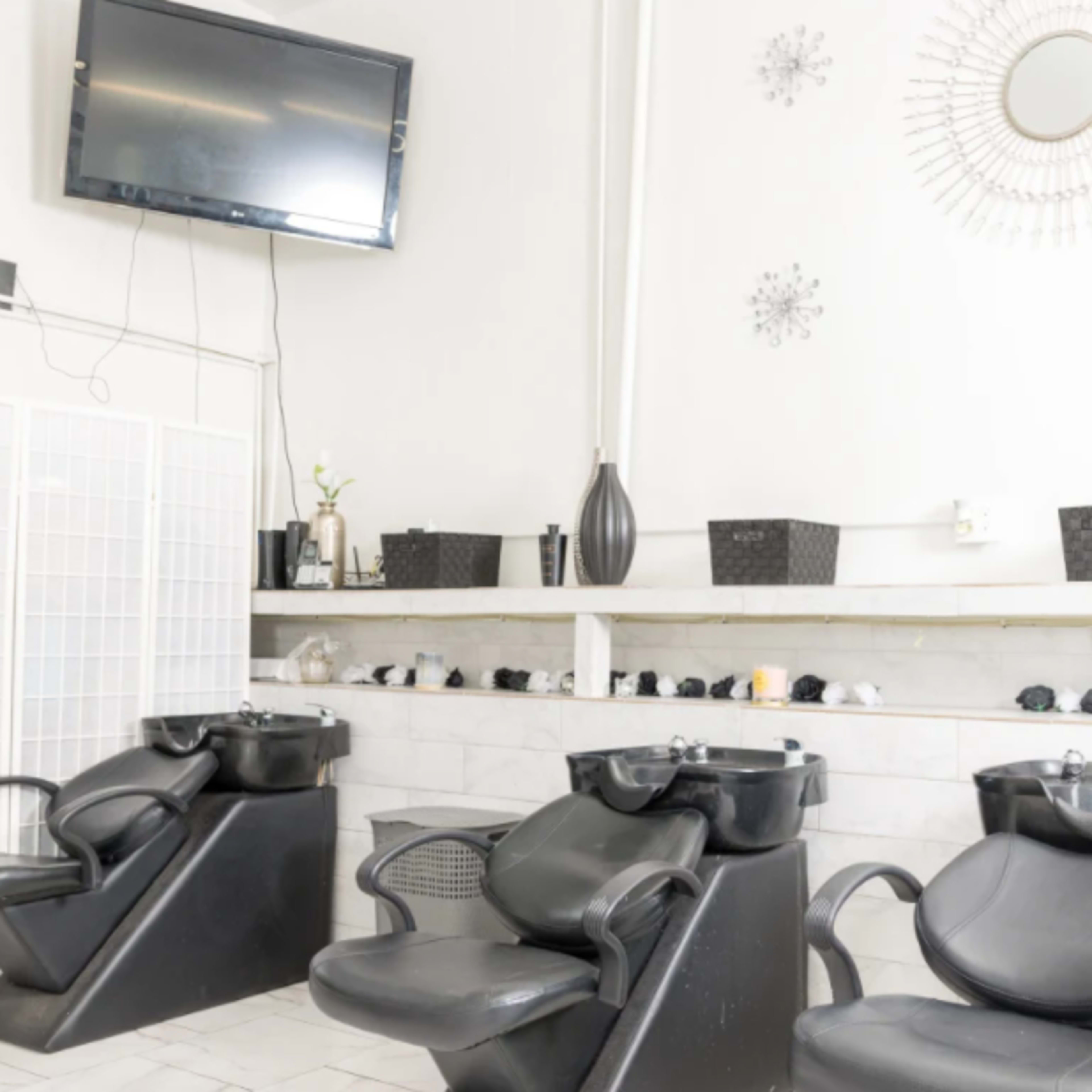 A hair salon interior with three black shampoo chairs, a wall-mounted television, and decorative items on shelves above.