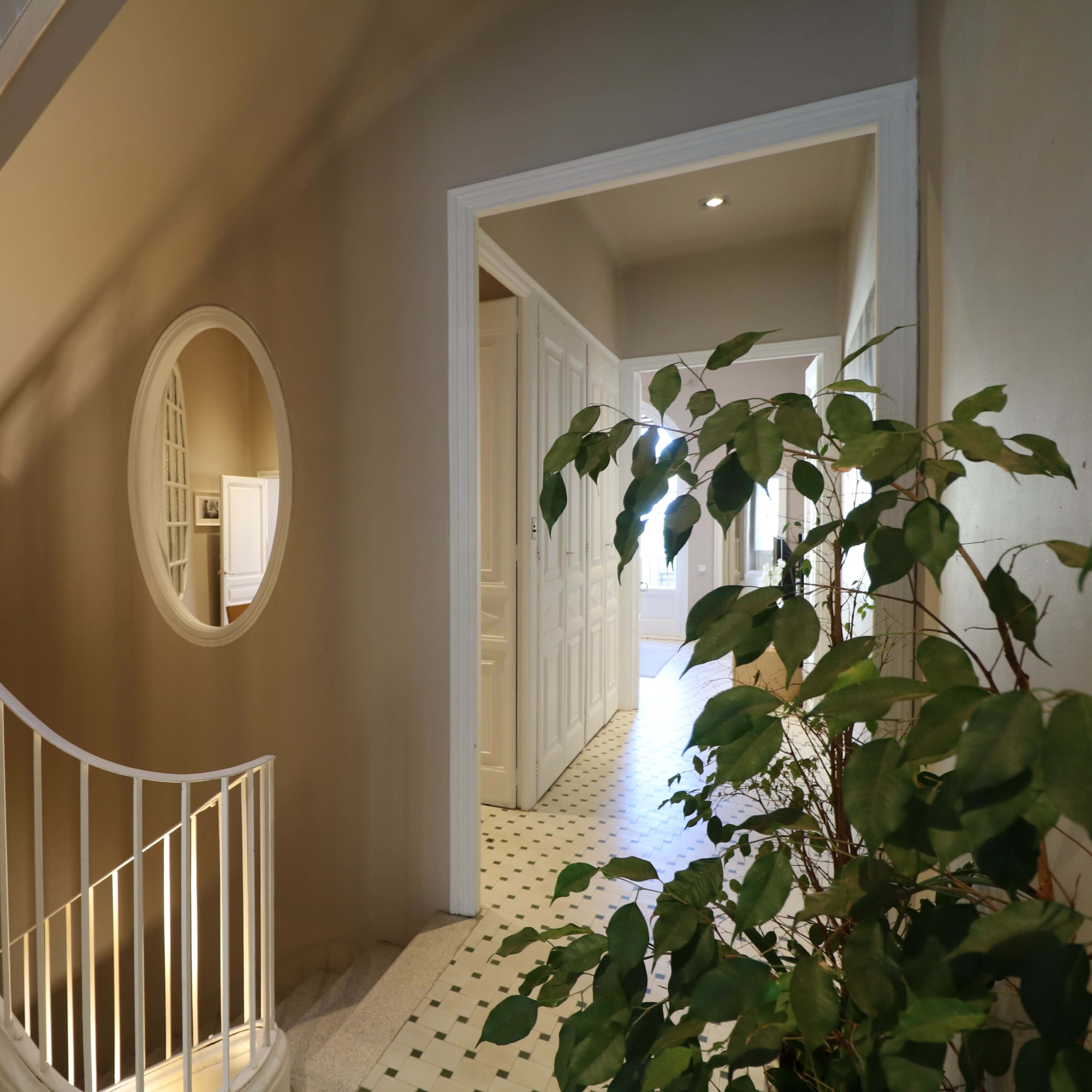 A staircase with a white railing leads to a hallway featuring an oval mirror and a floor with black and white tiles, complemented by a potted plant.