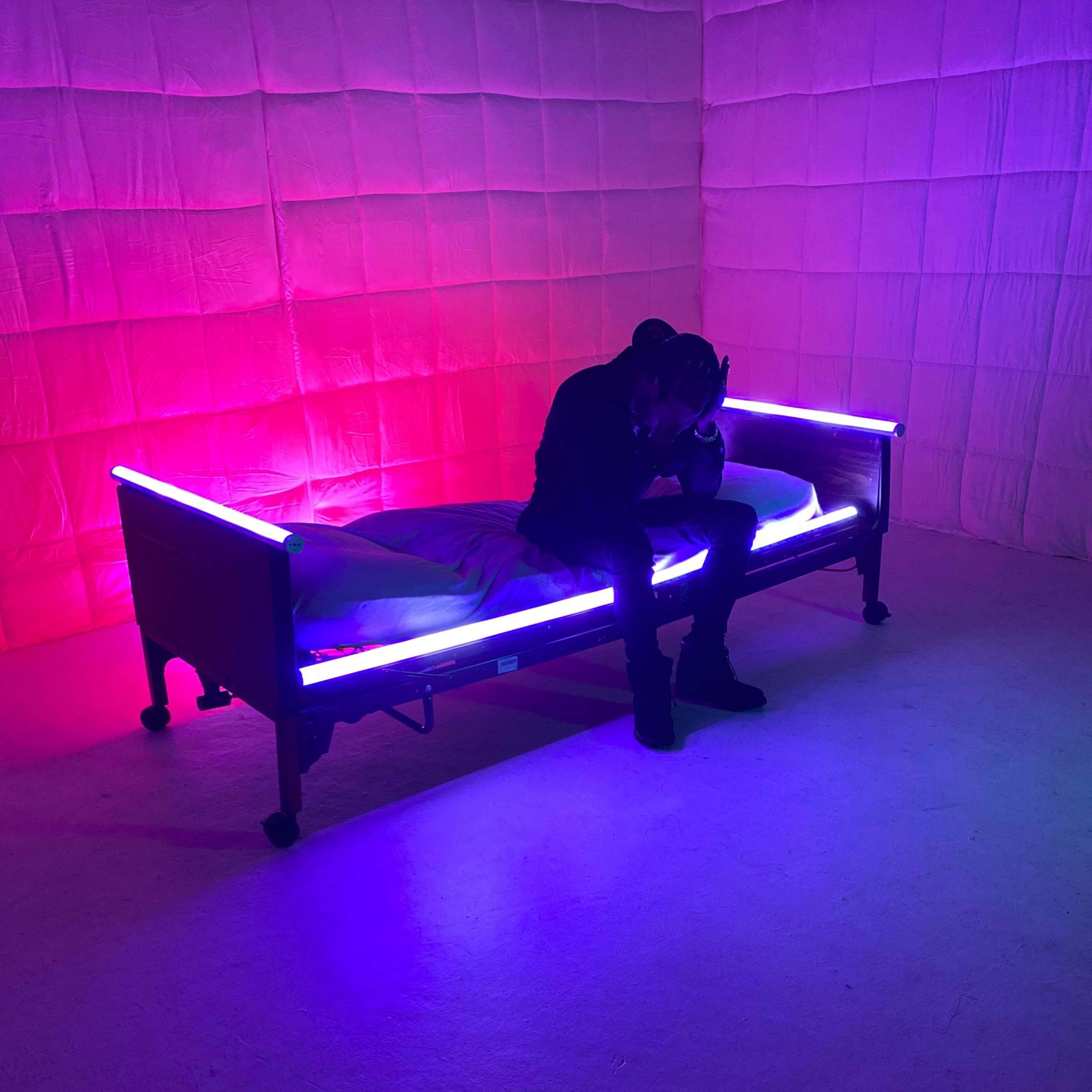 A person sits on a bed with glowing purple and pink lights in a room with textured walls.