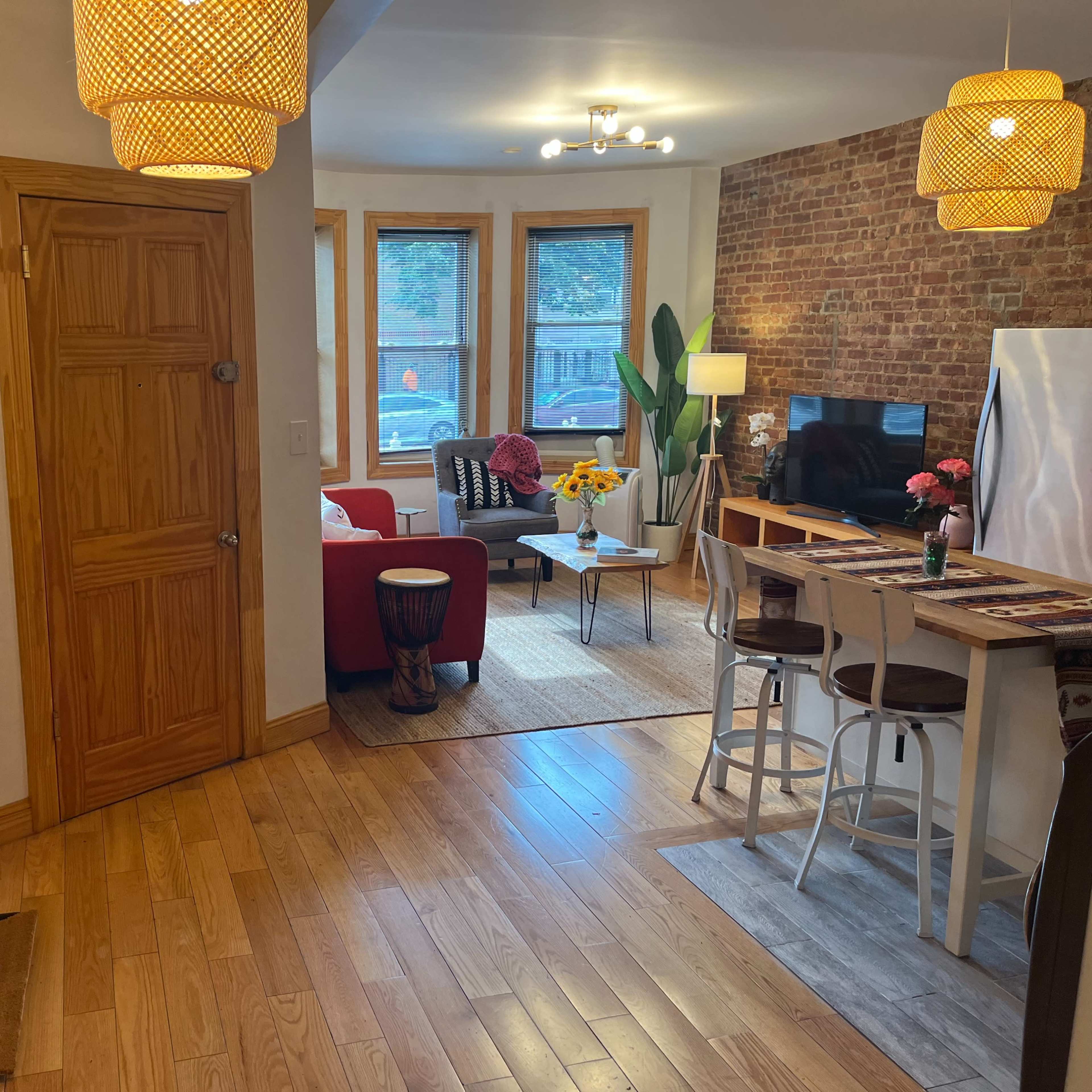 A spacious living area with a wooden floor, featuring a kitchen bar, a red chair, a television mounted on a brick wall, and decorative masks on the adjacent wall.