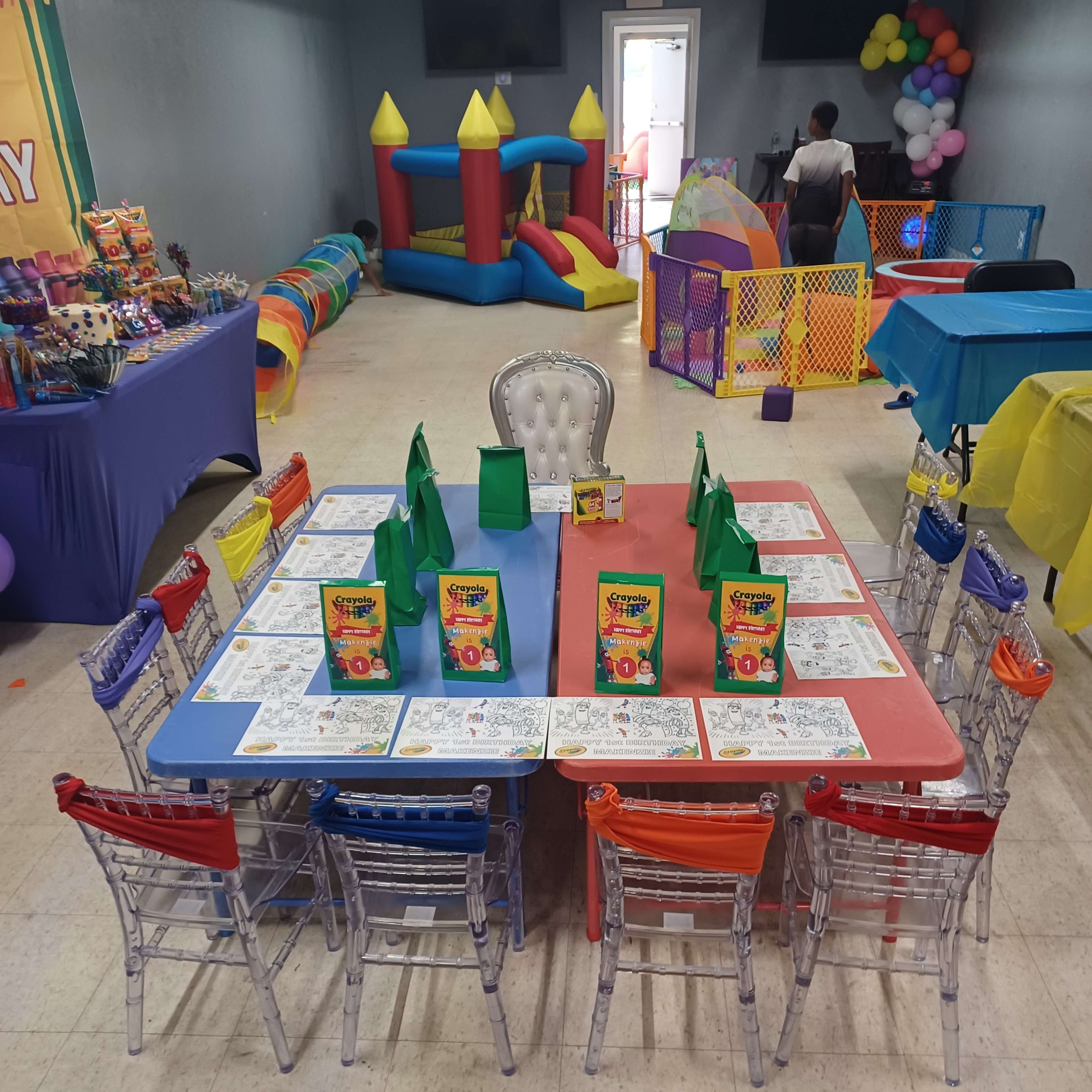 A brightly decorated party room features a large table set with coloring sheets and goody bags, surrounded by colorful chairs, while a bounce house and other play equipment are visible in the background.
