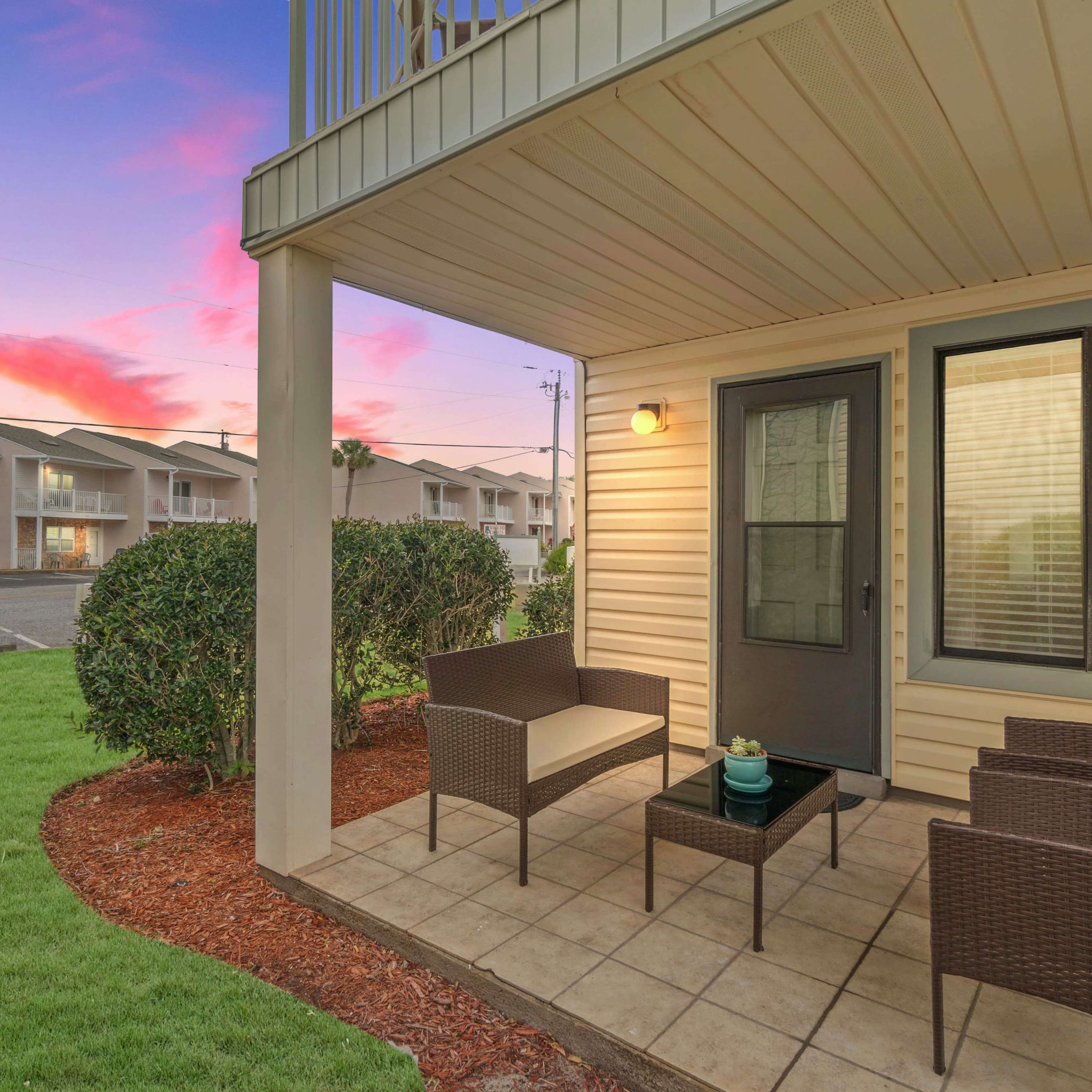 A patio area features outdoor seating with wicker chairs and a table, surrounded by landscaped grass and bushes, set against a colorful sunset backdrop.