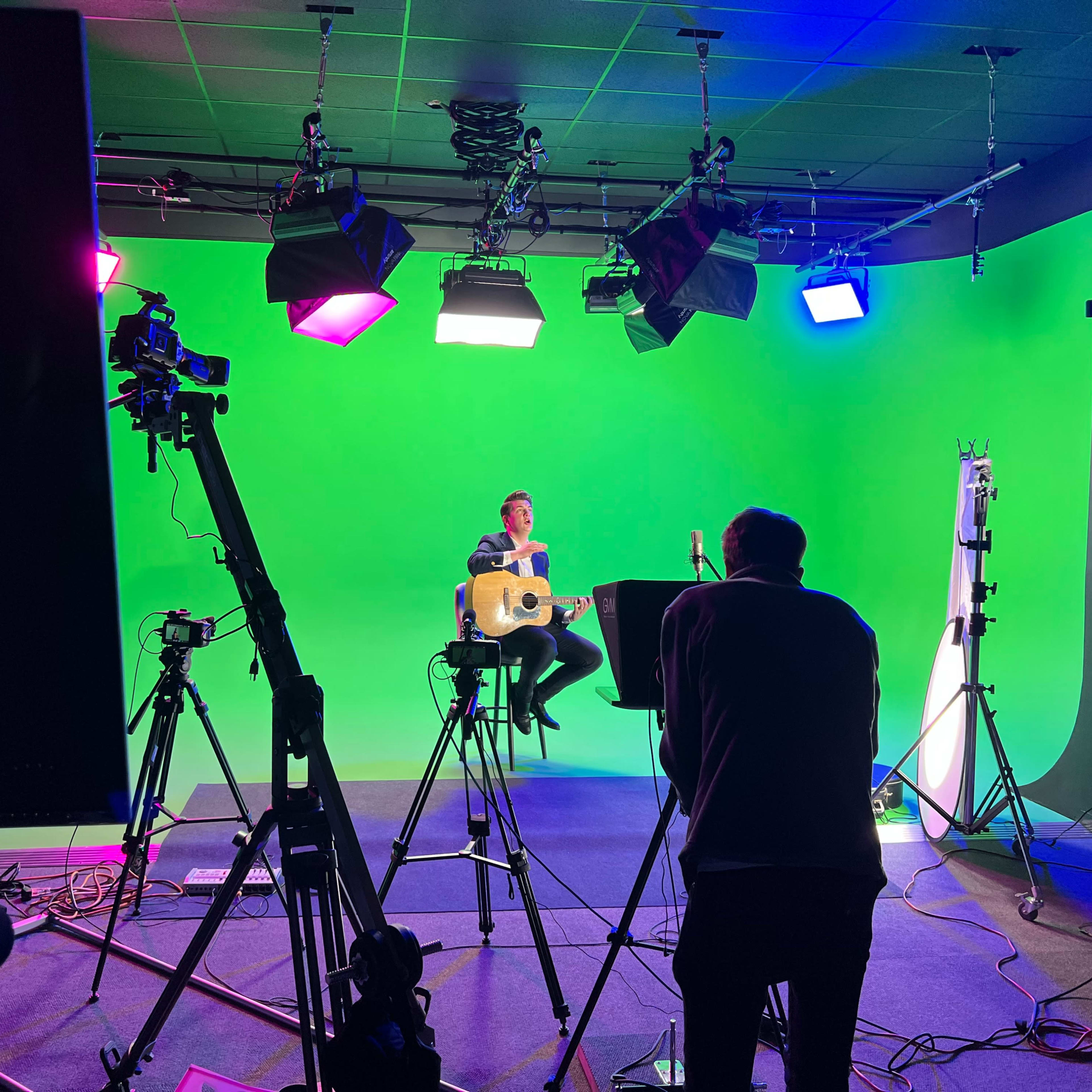 A musician sits on a stool with a guitar in front of a green screen, surrounded by multiple cameras and studio lights.