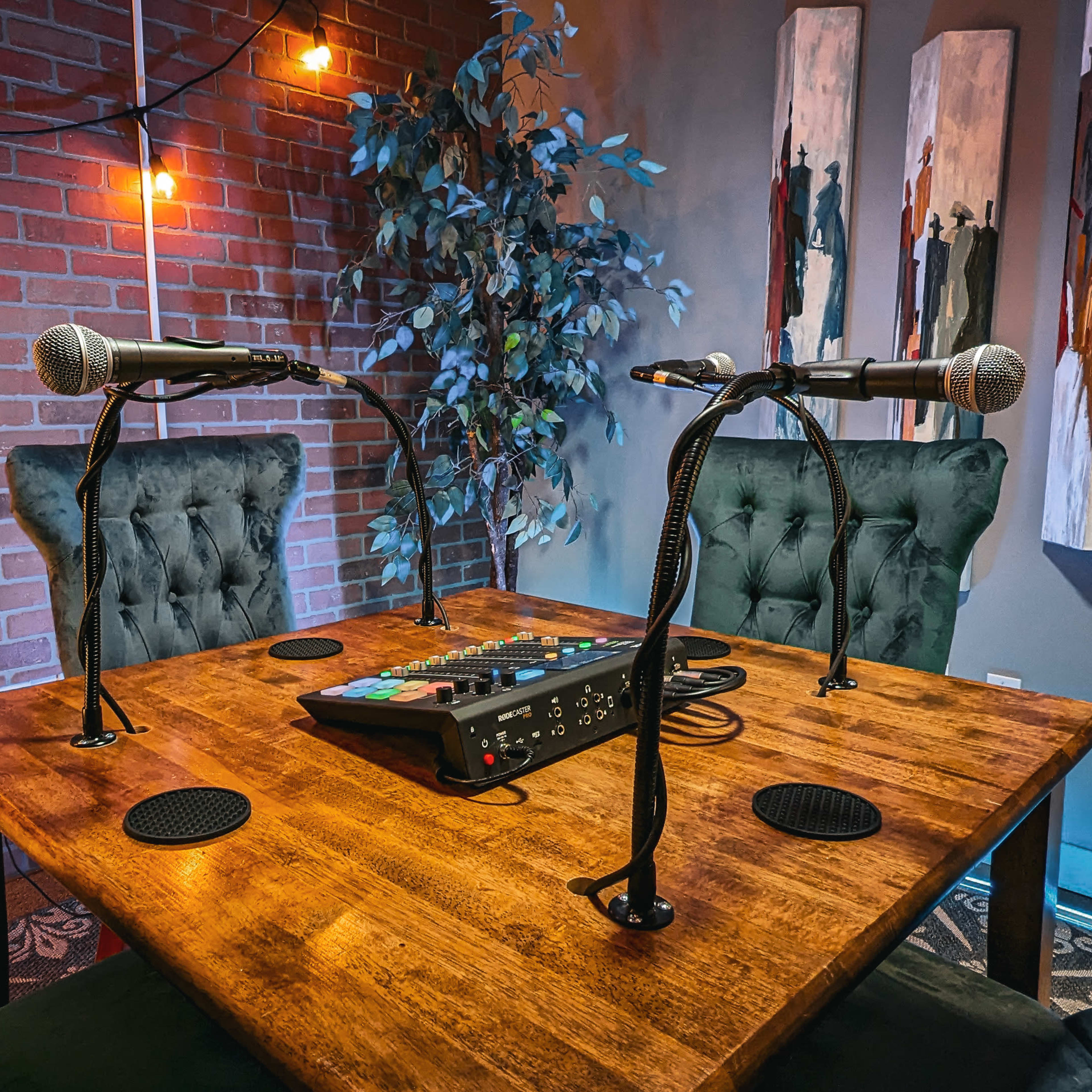 A podcast recording setup with two microphones on a wooden table, surrounded by green chairs and decorated walls featuring artwork and string lights.