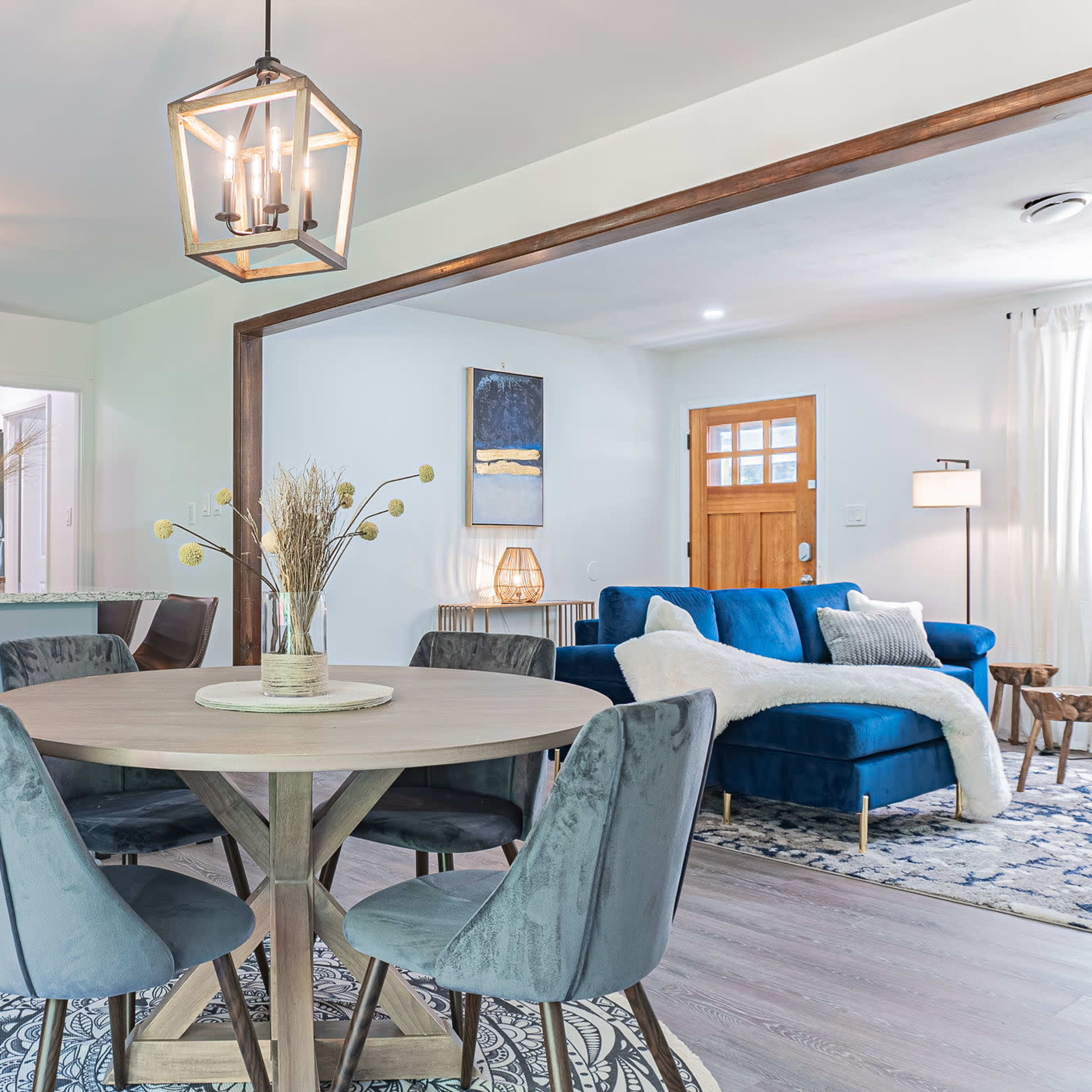 A modern kitchen and dining area with a circular wooden table surrounded by grey chairs, leading into a cozy living room with a blue couch and bright lighting.