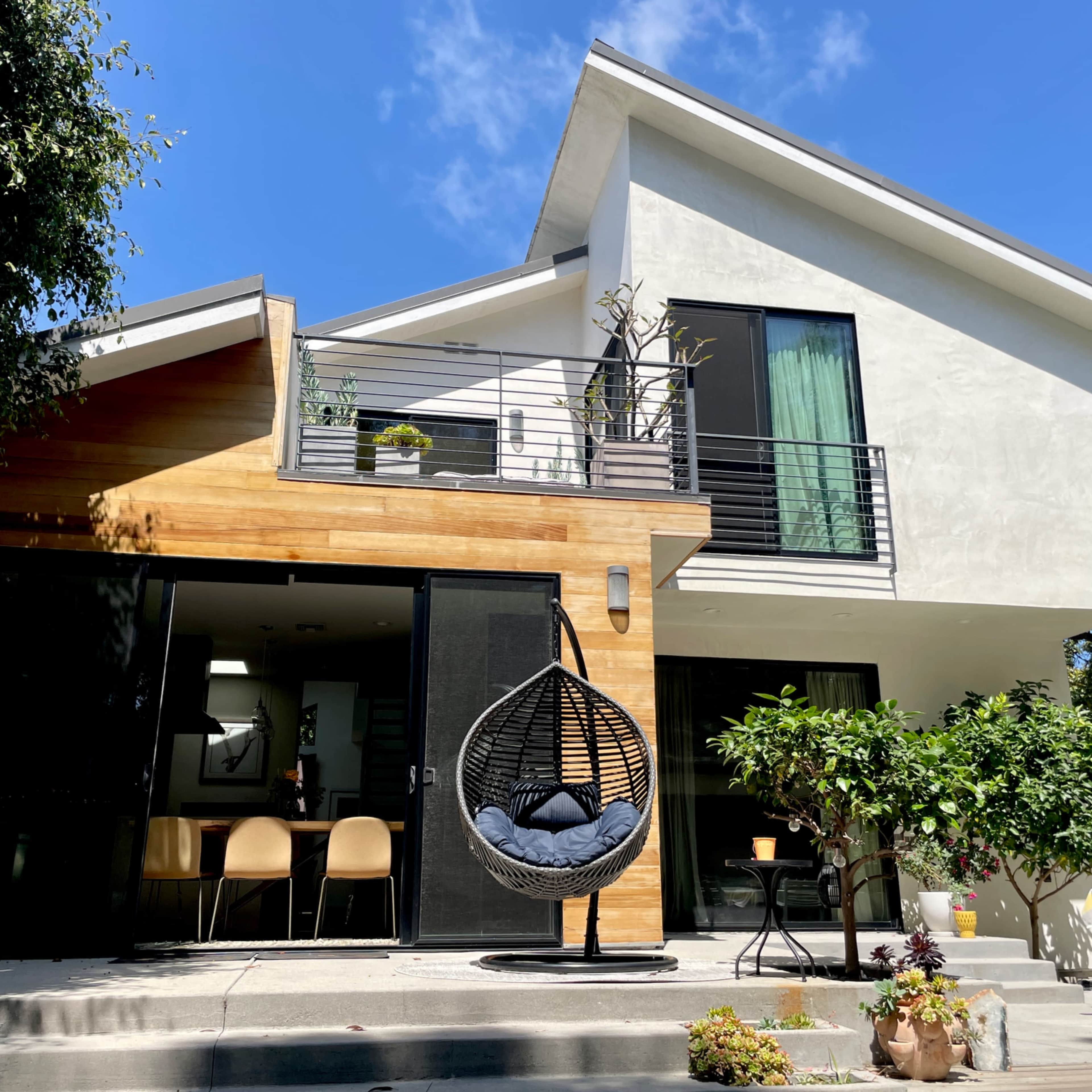 A modern two-story house features a combination of wood and white exterior, with a hanging chair on the patio and large windows.