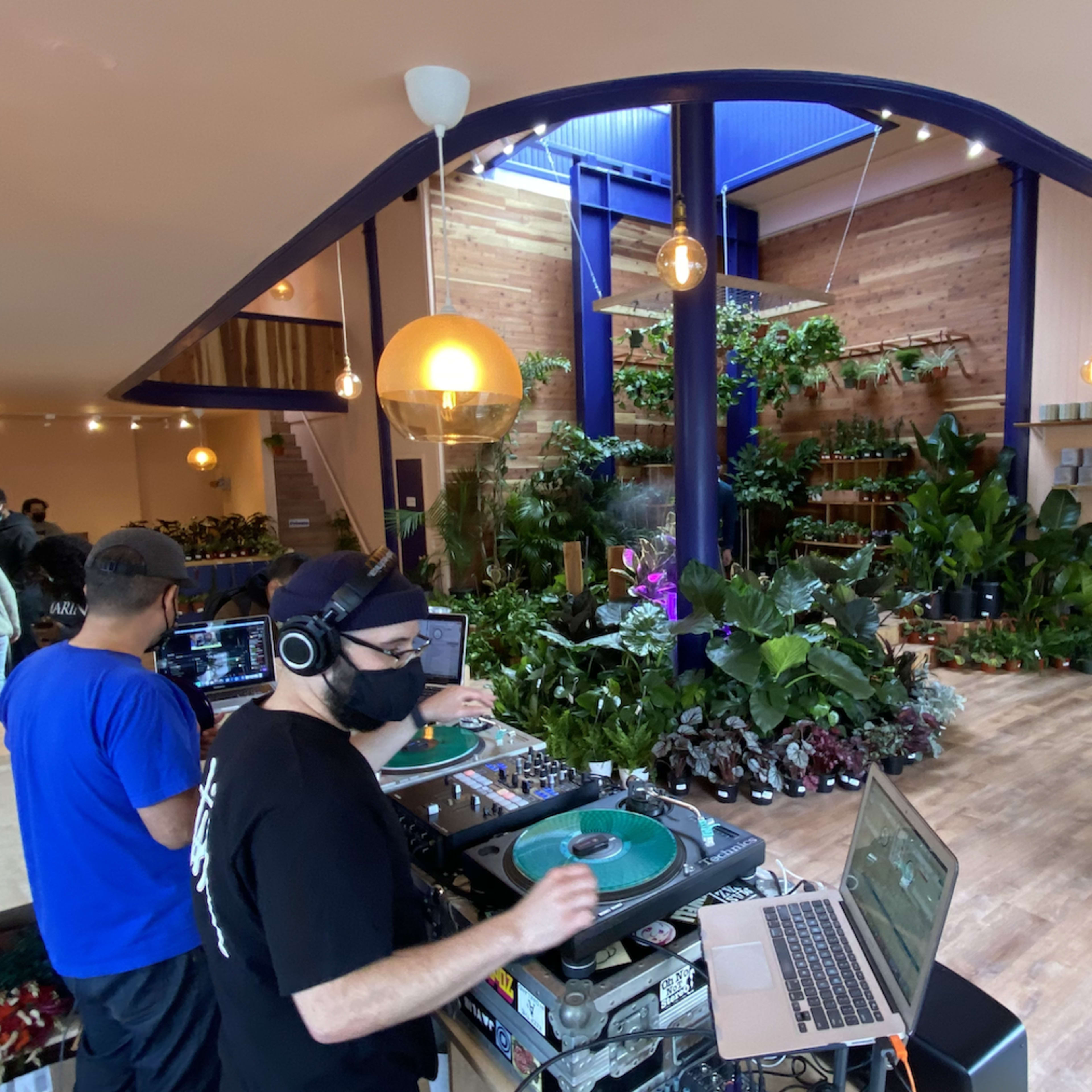 A DJ stands behind turntables in a plant-filled store while customers browse the greenery.