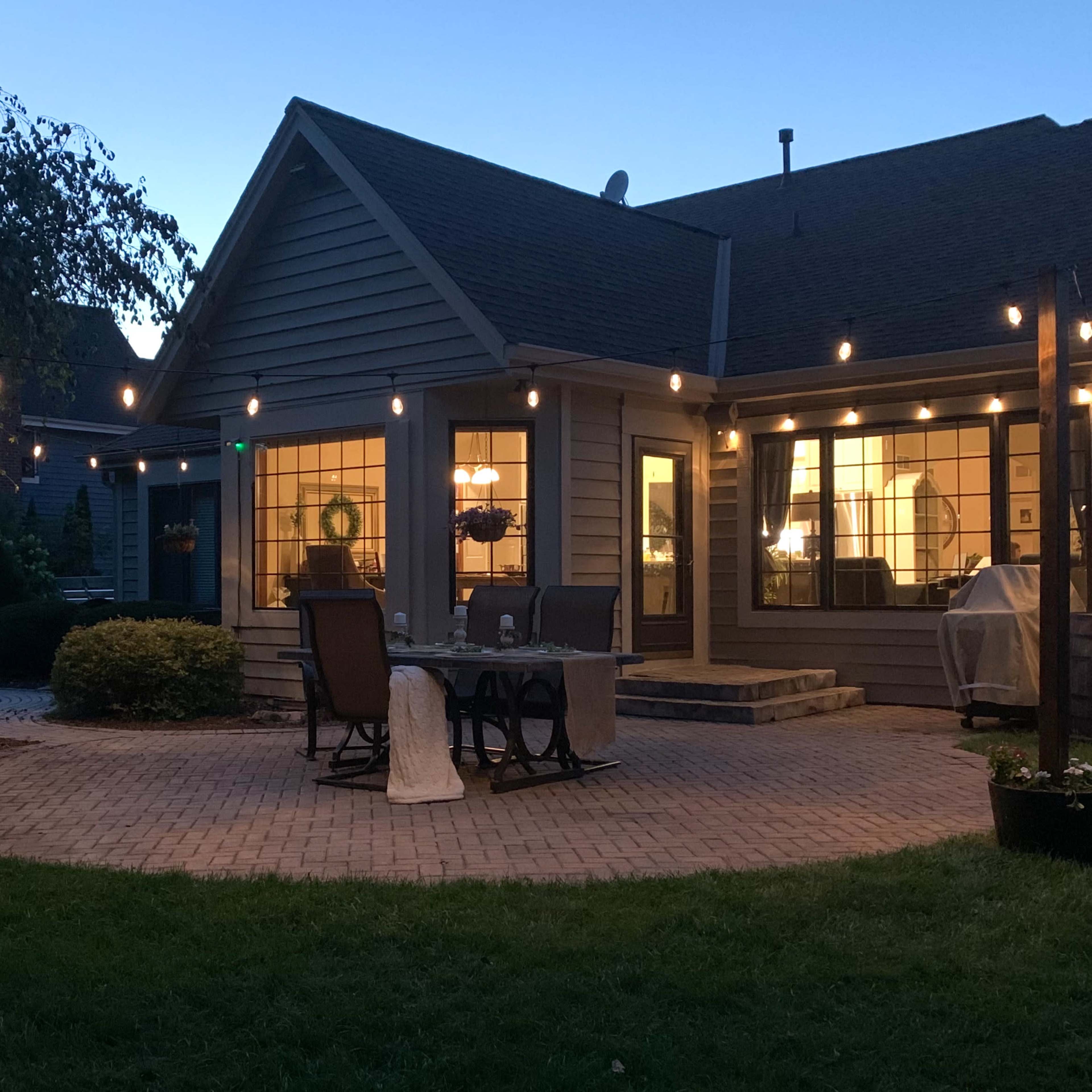 A patio is set up behind a house, illuminated by string lights, with outdoor furniture arranged on a brick surface.