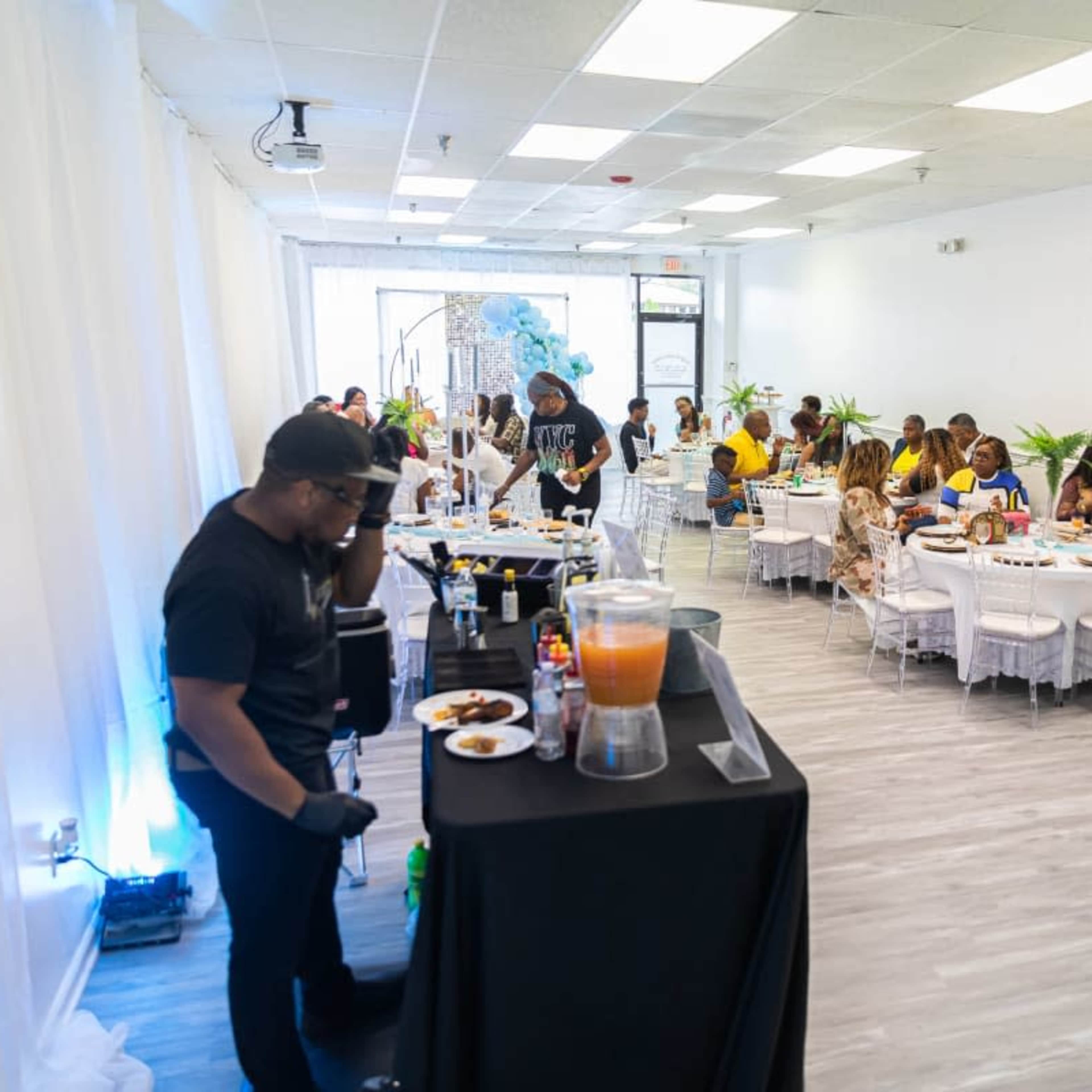 A spacious event room is set up with tables filled with guests enjoying a meal, while a person at a buffet table prepares food.