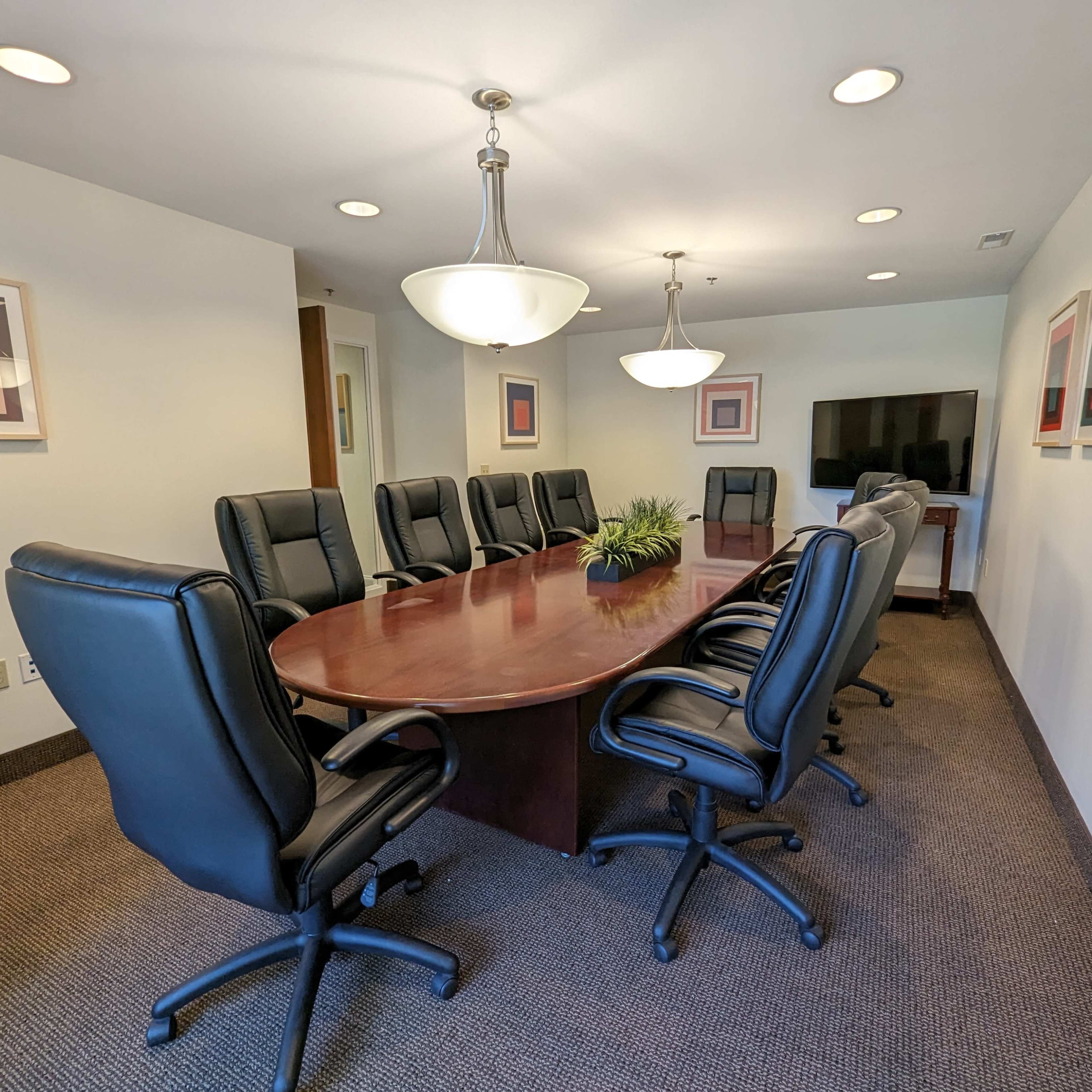 A formal meeting room features a large oval table surrounded by eight black leather chairs, with two additional chairs at one end and modern artwork on the walls.