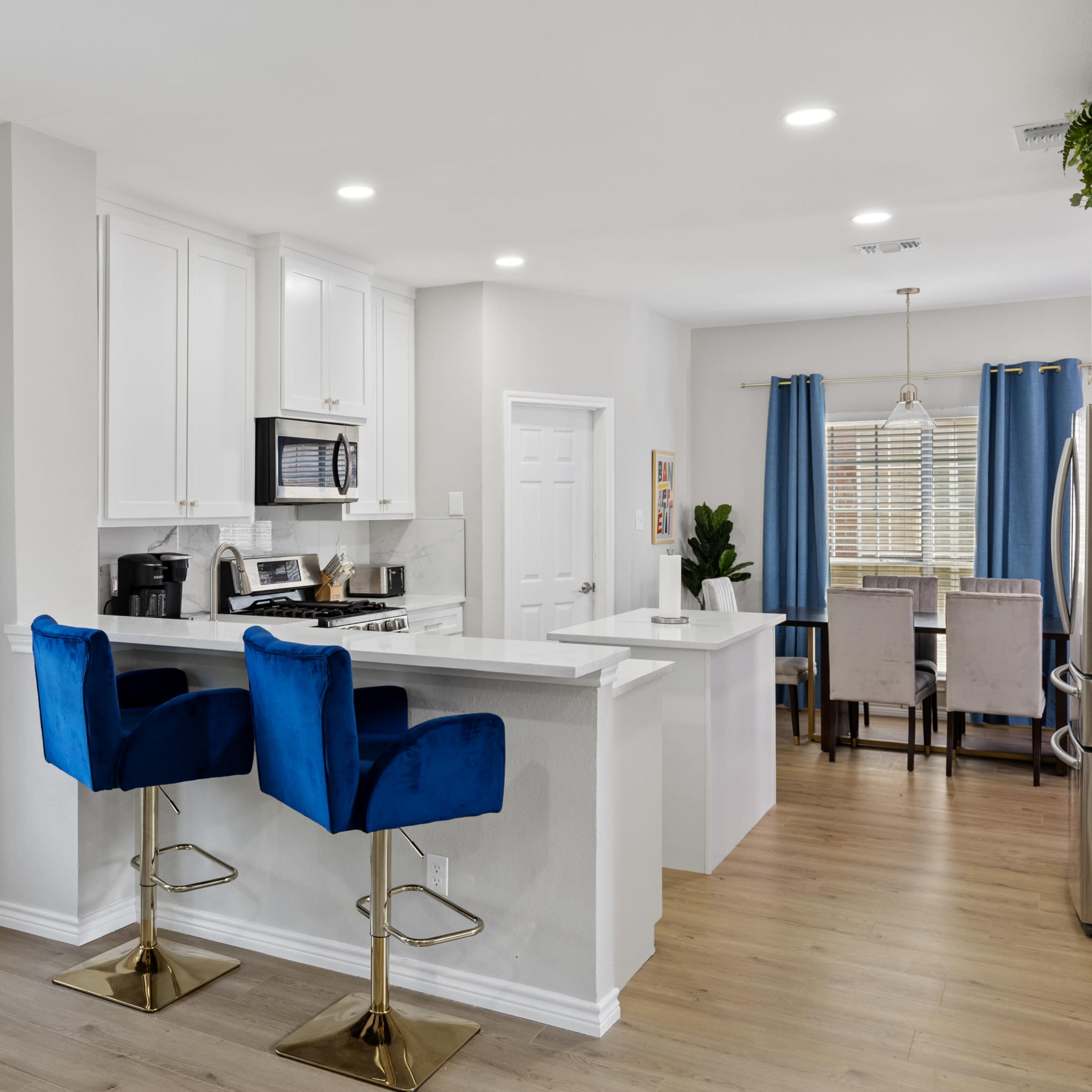 A modern kitchen features white cabinetry, stainless steel appliances, and blue bar stools at a counter, with a dining area visible in the background.