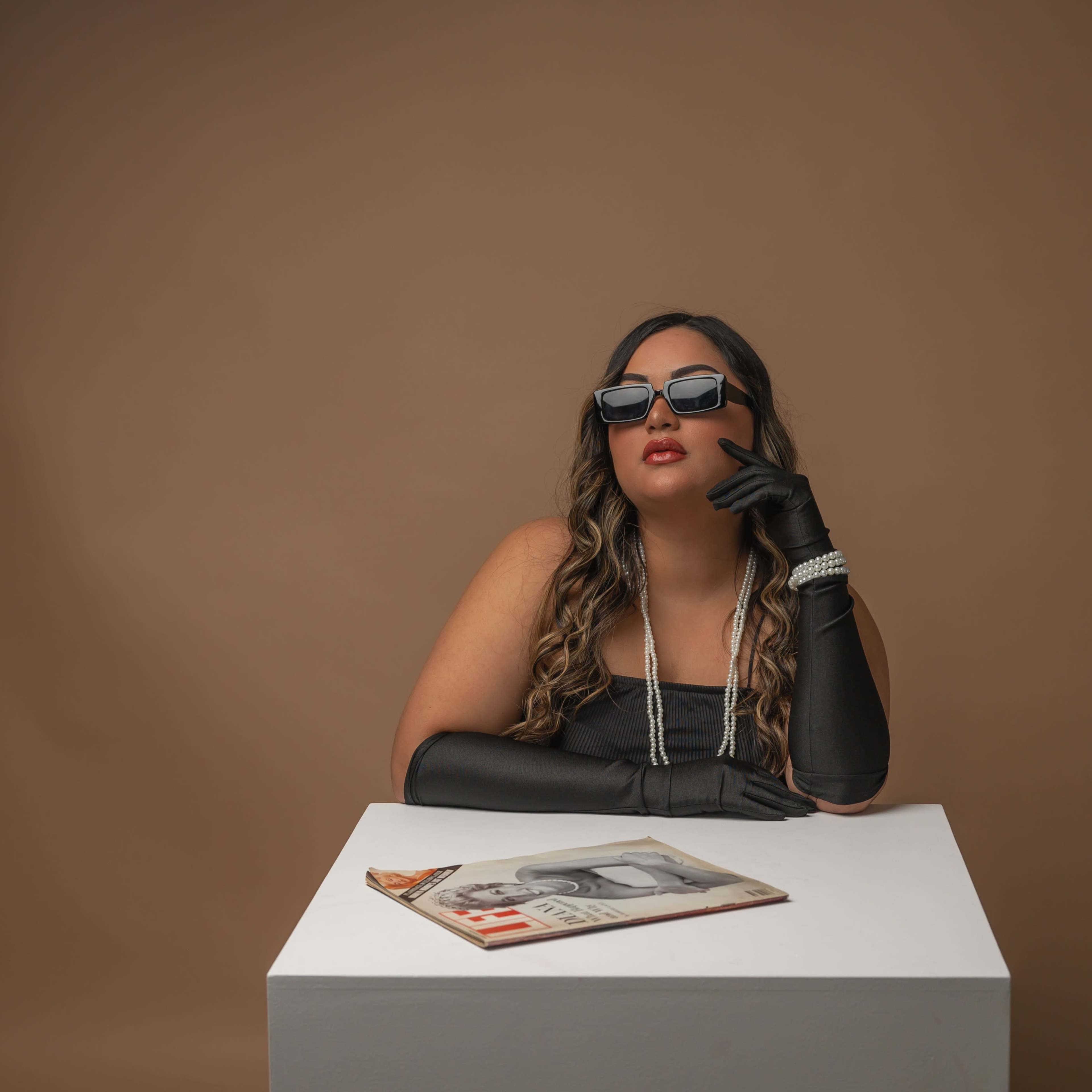 A woman in black sunglasses and long gloves sits at a white table with a magazine beside her, against a brown backdrop.