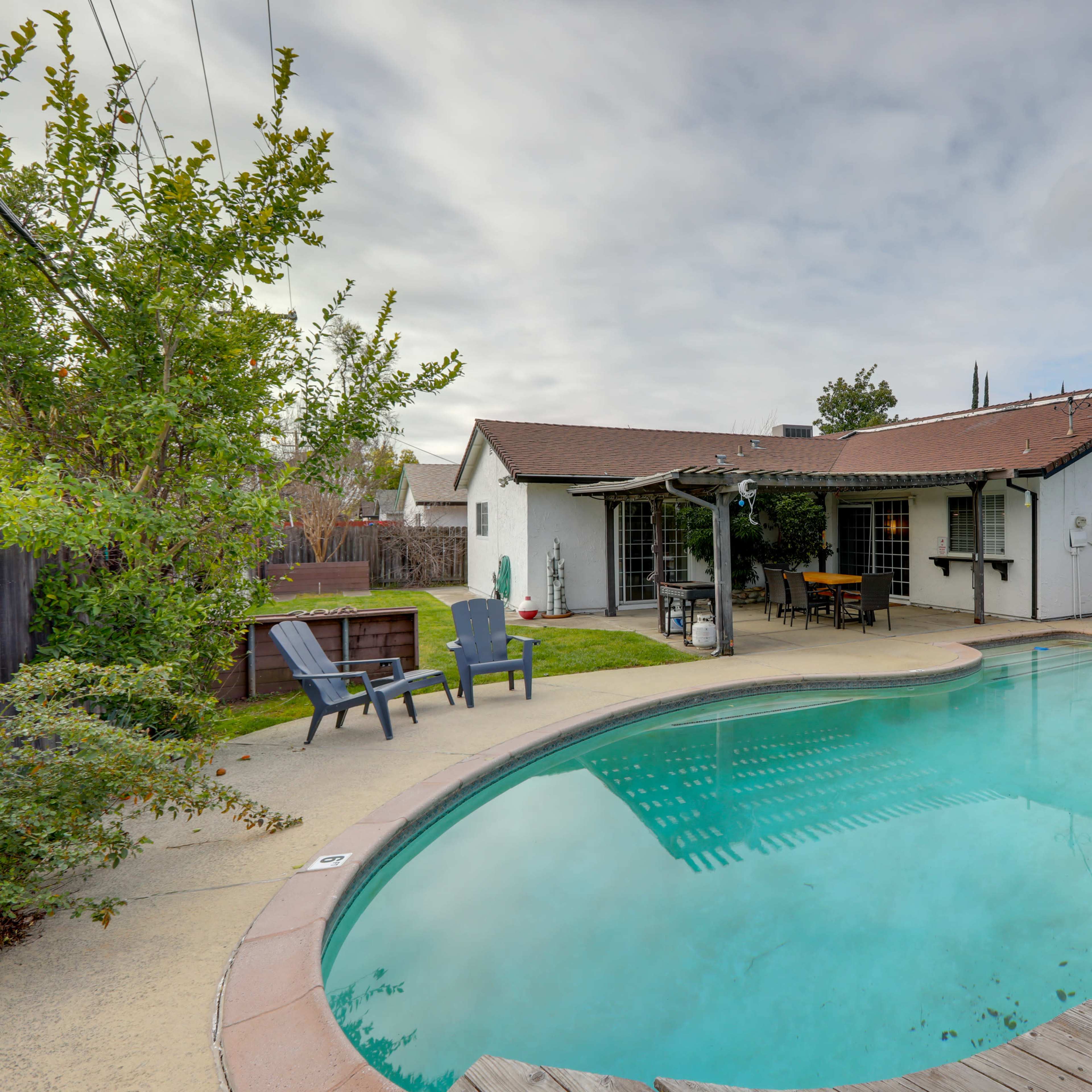 A backyard scene features a swimming pool surrounded by a wooden deck and a single-story house with a patio area.