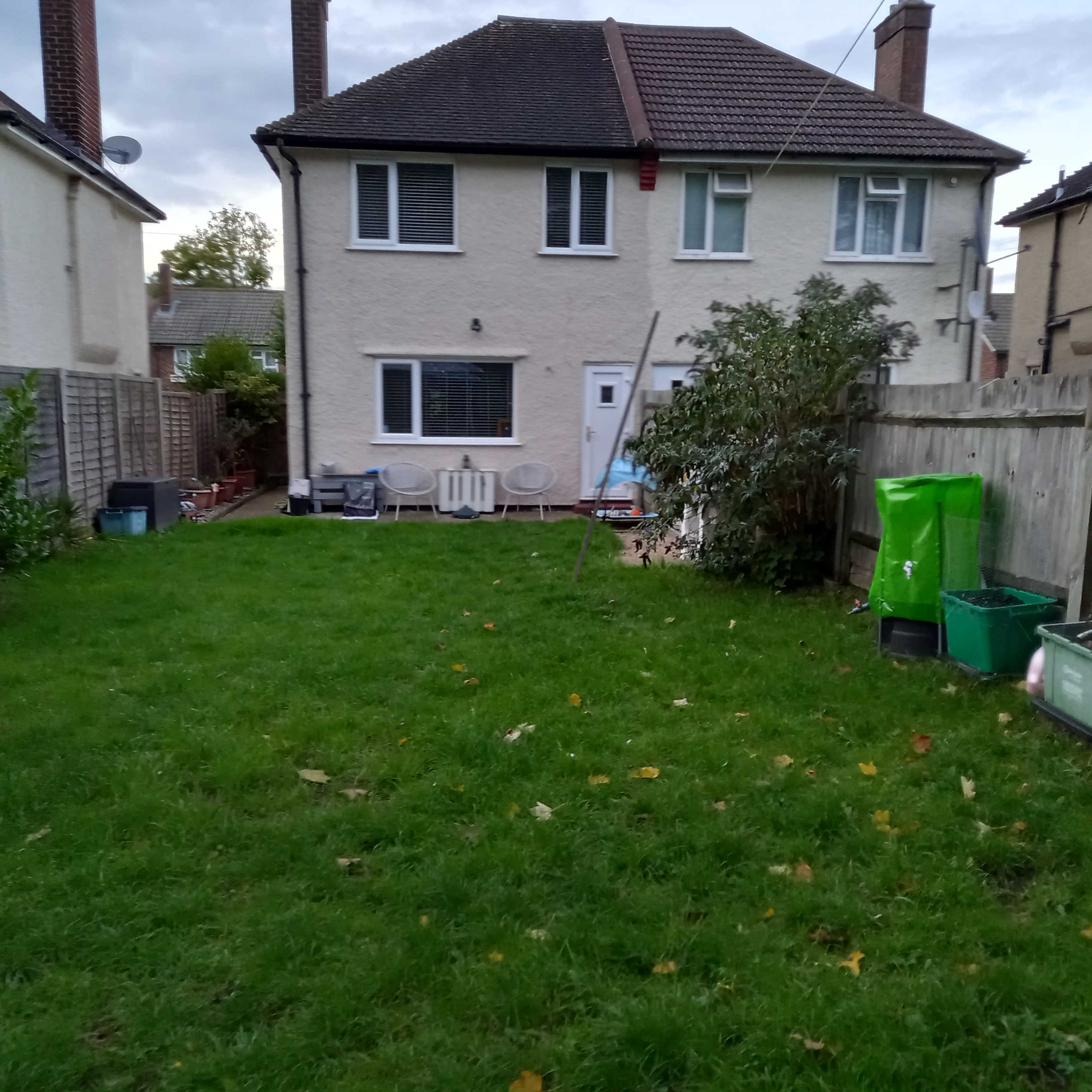 A grassy backyard with a two-story beige house, green garbage bins, and a fence surrounding the area.