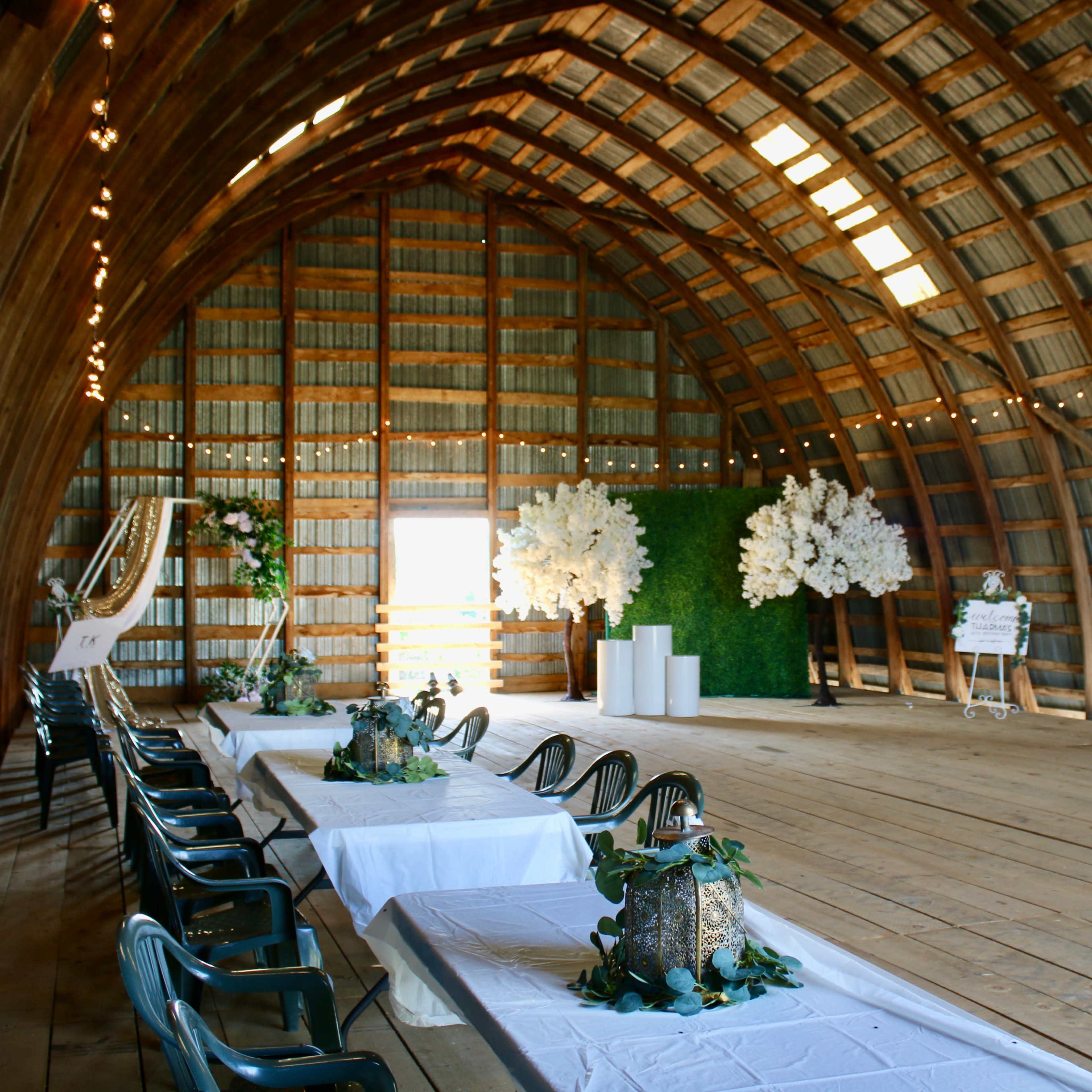A spacious wooden barn interior set up for an event, featuring a long table with decorations, chairs, and a decorative backdrop of white flowers and greenery.