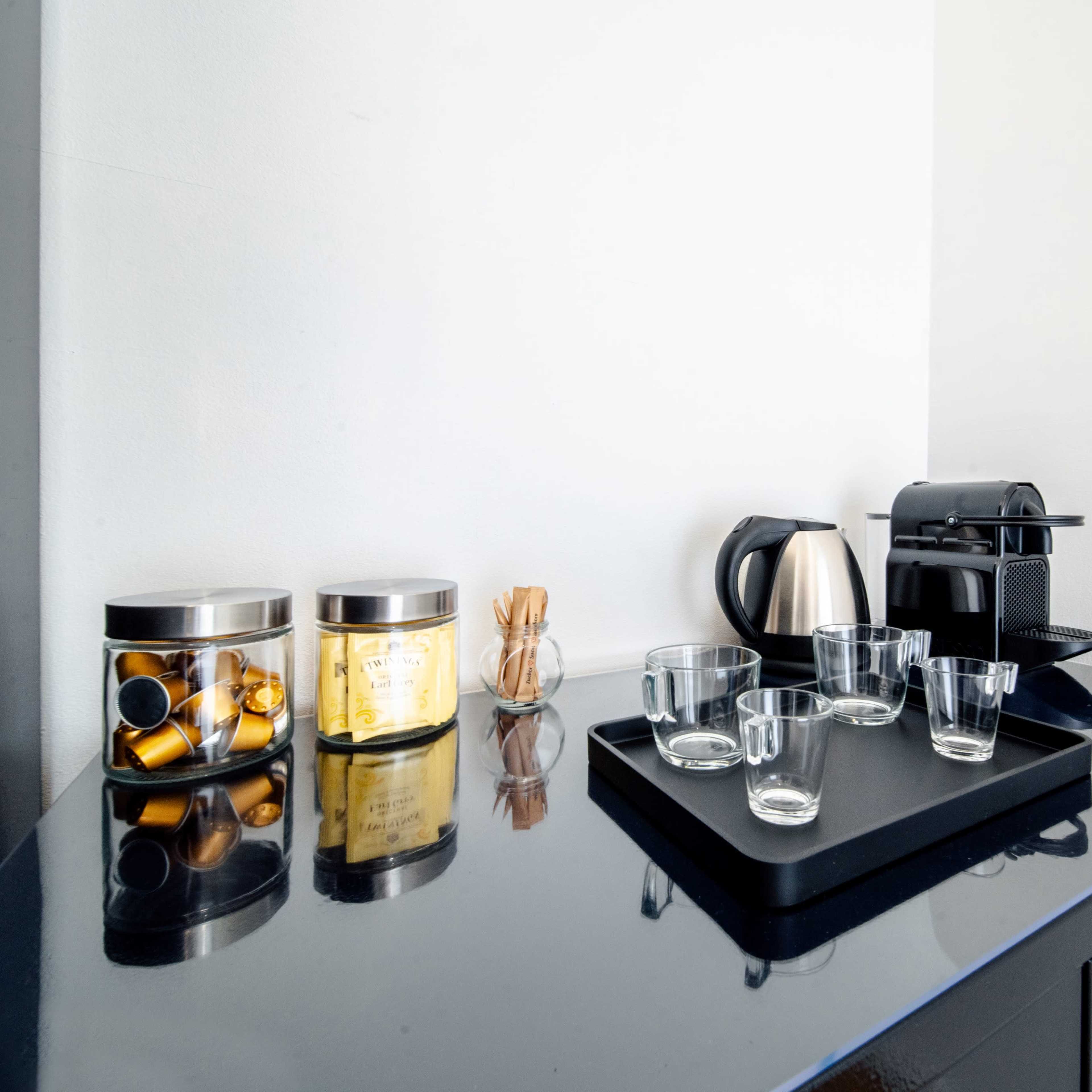 A coffee station is set up on a countertop, featuring a coffee machine, a kettle, several glass cups, and jars with coffee capsules and stirrers.