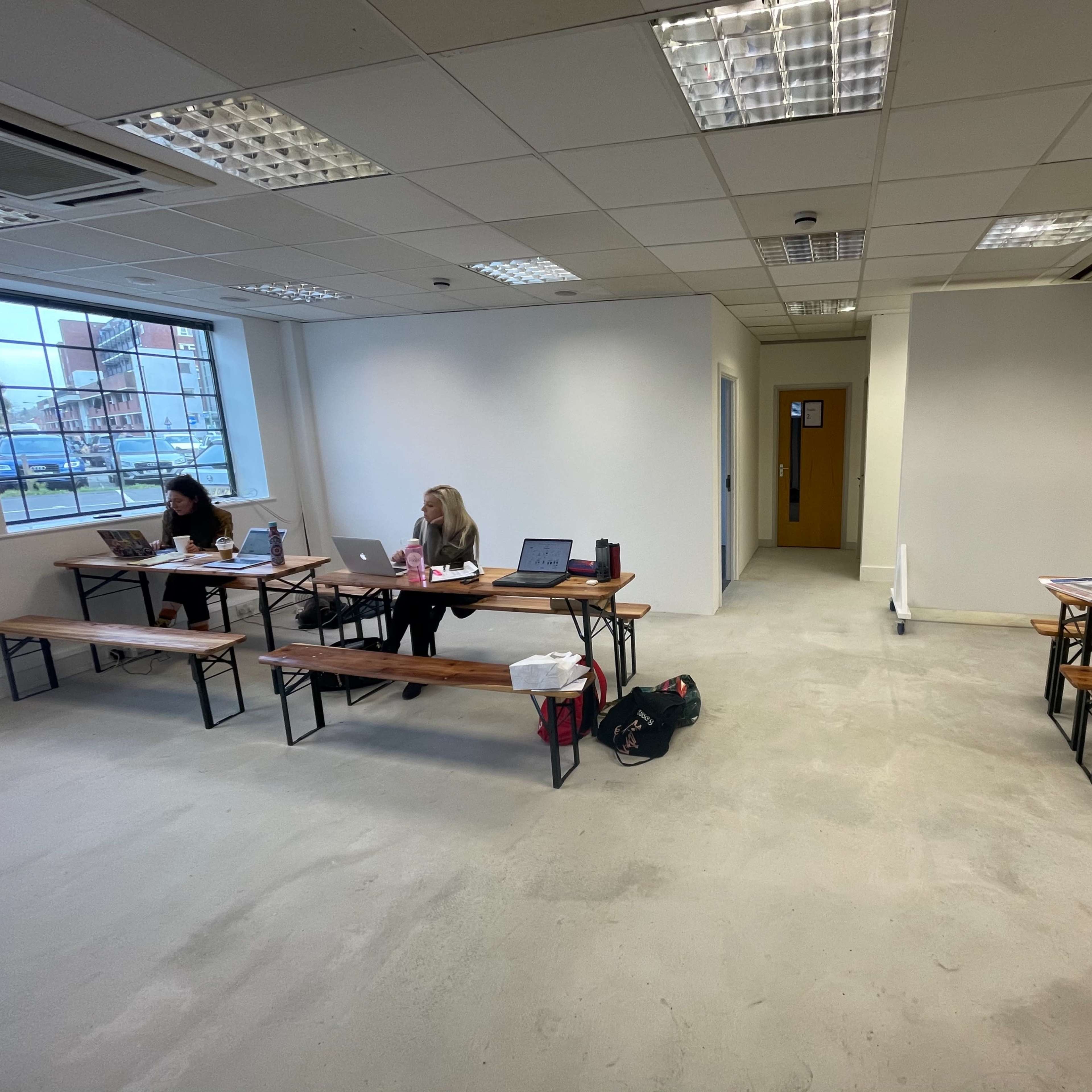 Two women sit at wooden tables with laptops in a sparsely furnished classroom featuring concrete floors and large windows.
