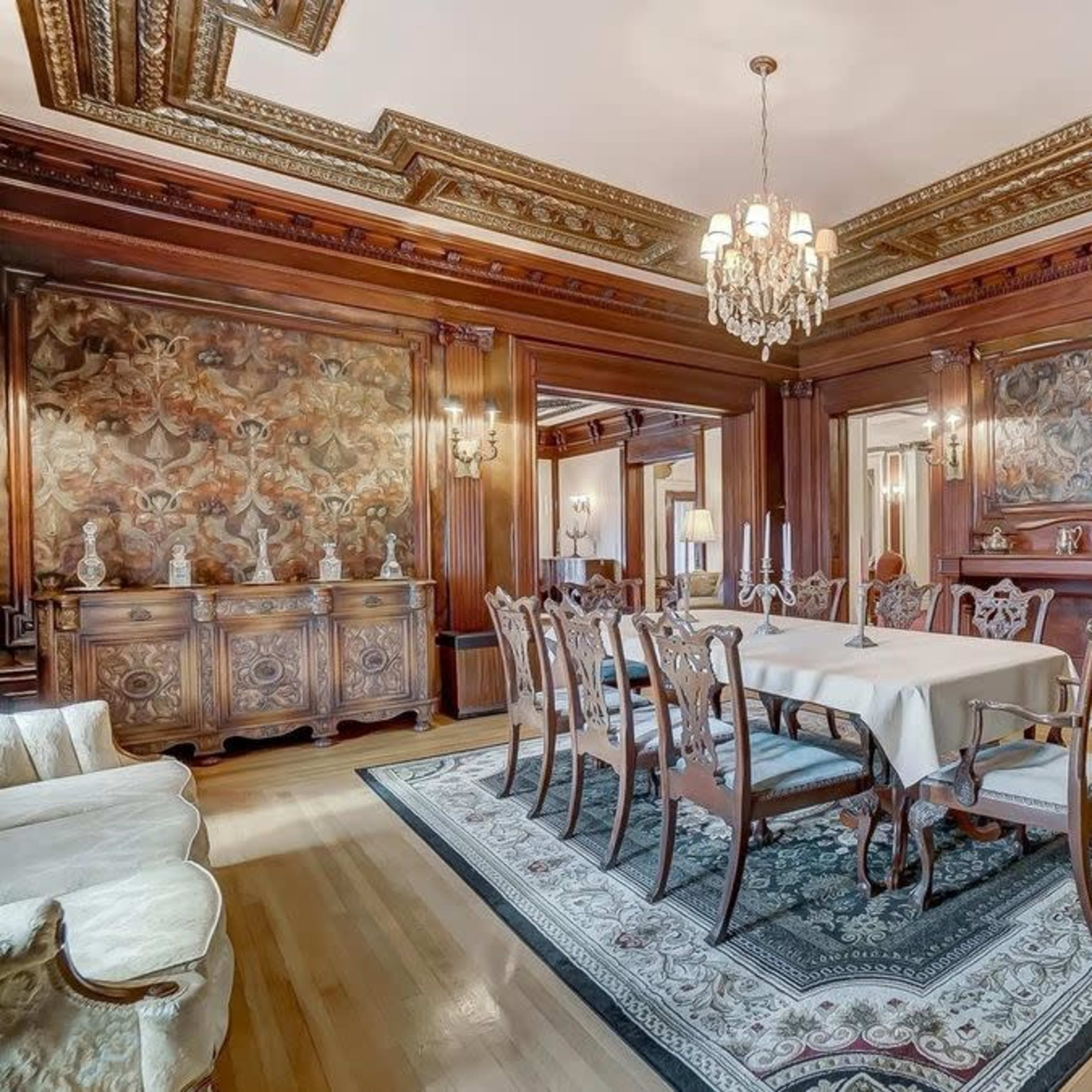 An elegant dining room with a large table surrounded by ornate chairs, featuring detailed wood paneling and a chandelier overhead.