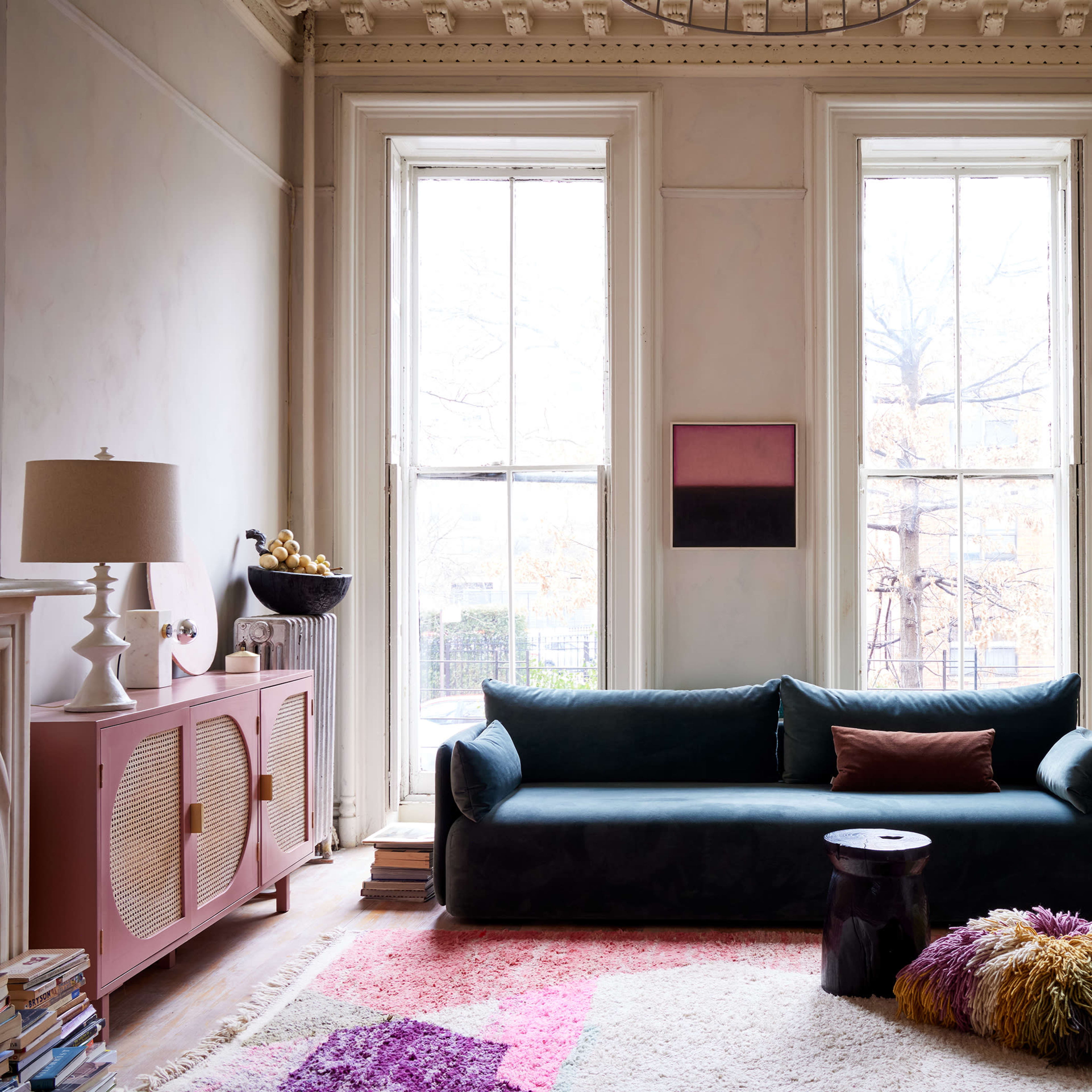 The living room features a dark green sofa, a pink sideboard, large windows with natural light, and decor that includes plants and artwork on the walls.