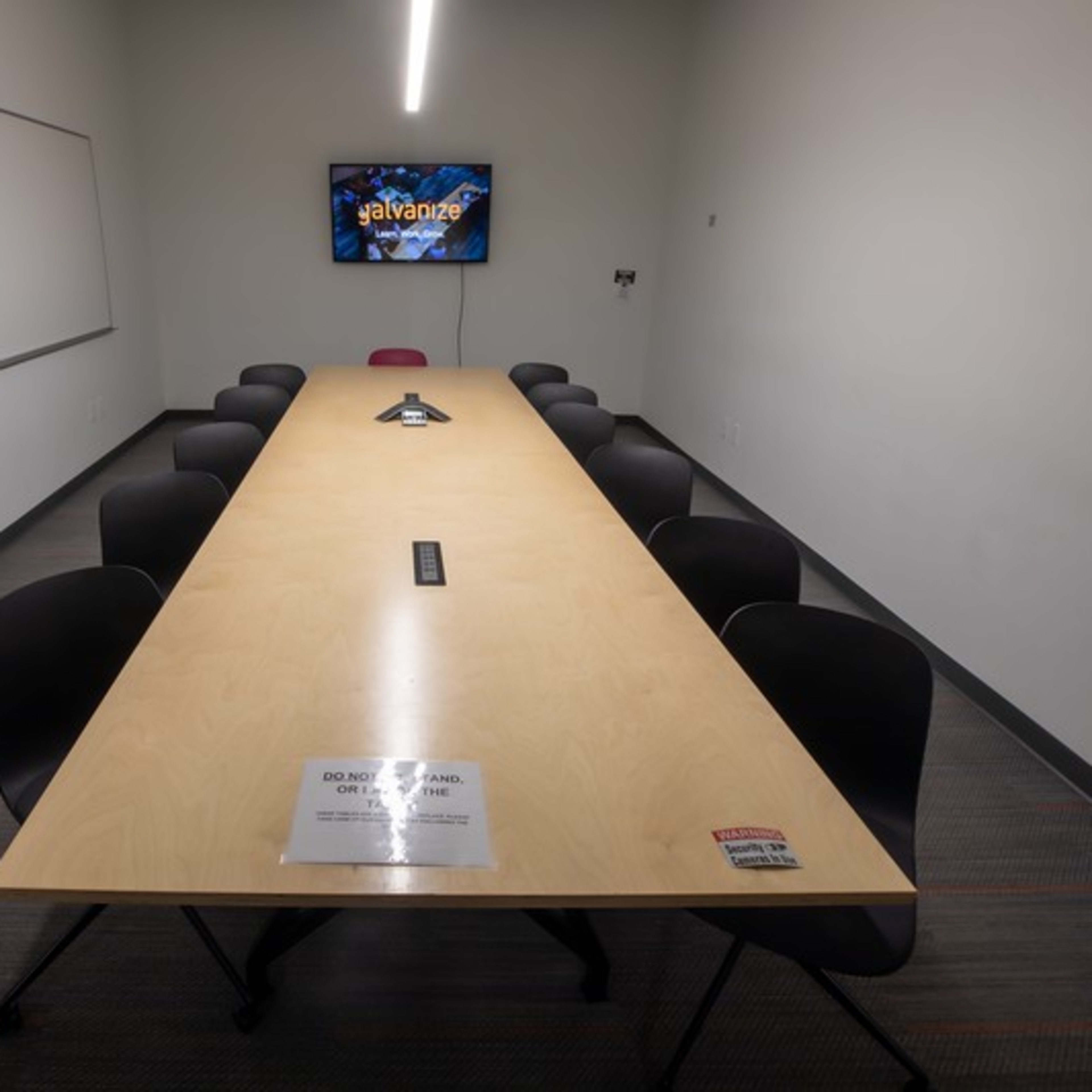 A long wooden conference table with eight black chairs is situated in a meeting room, featuring a television mounted on the wall and a sign on the table.