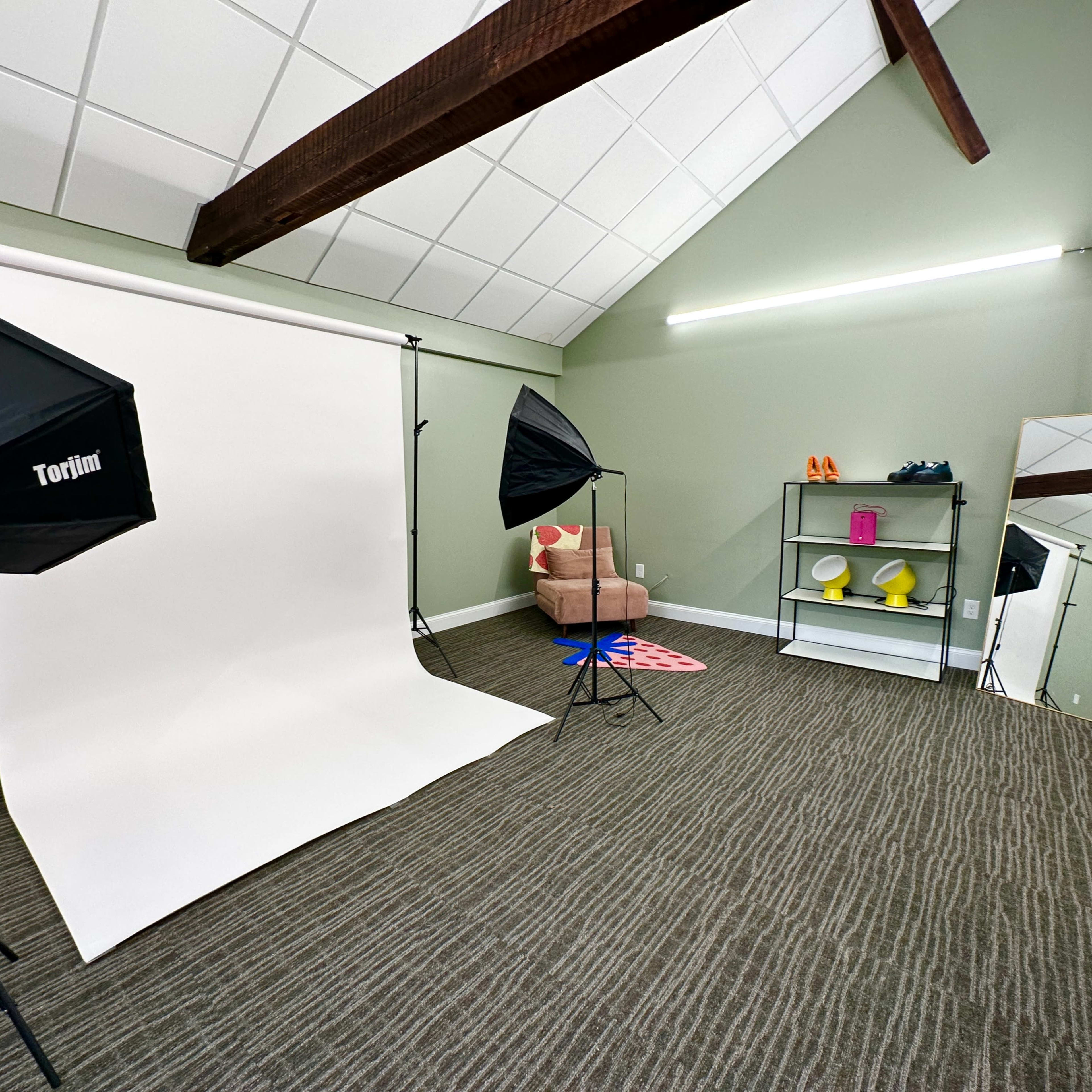 A small studio space with a white backdrop, lighting equipment, and a pink chair positioned beside a mirror and shelves with colorful props.