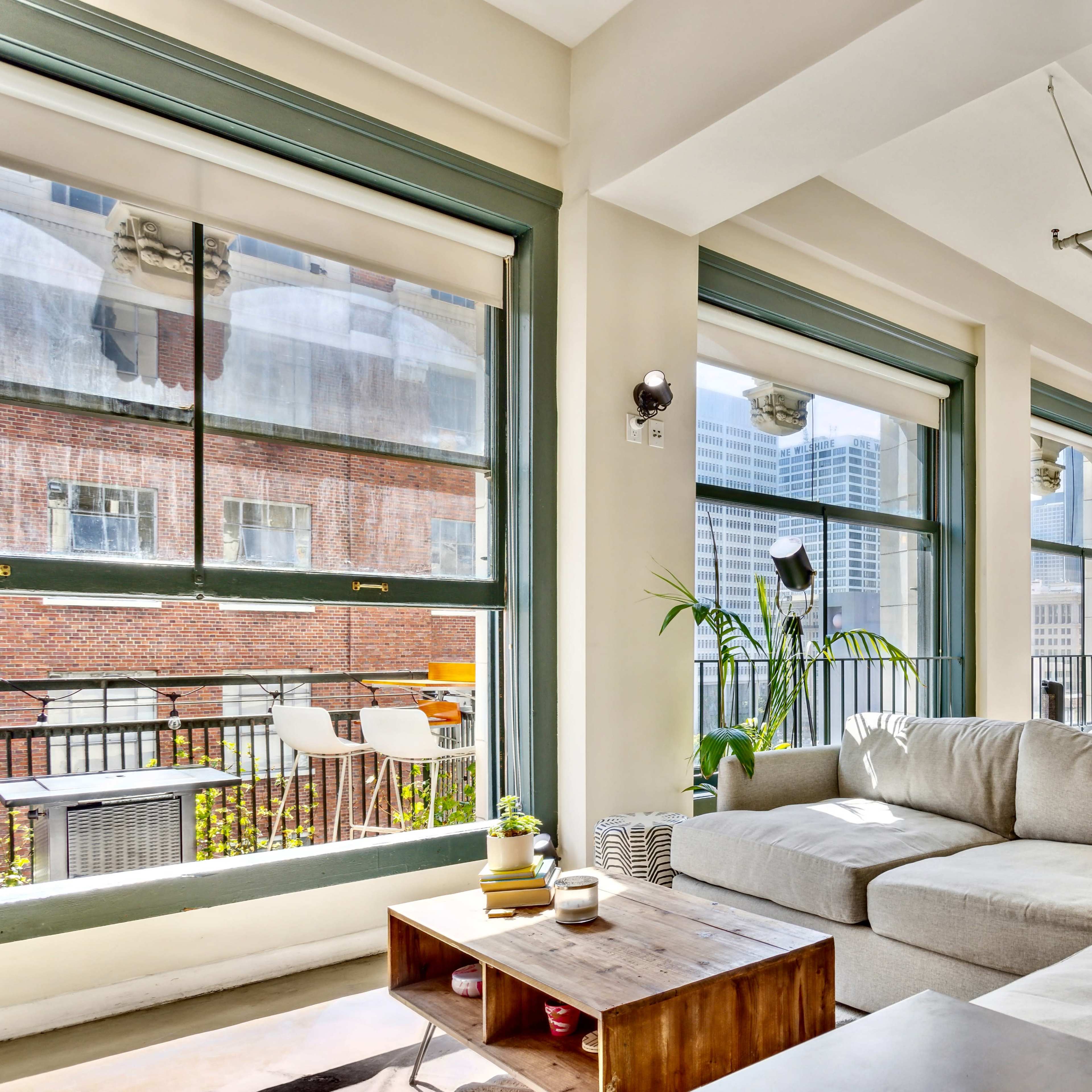 A bright, modern living room features a large sectional couch, a wooden coffee table, and expansive windows that offer views of a cityscape.