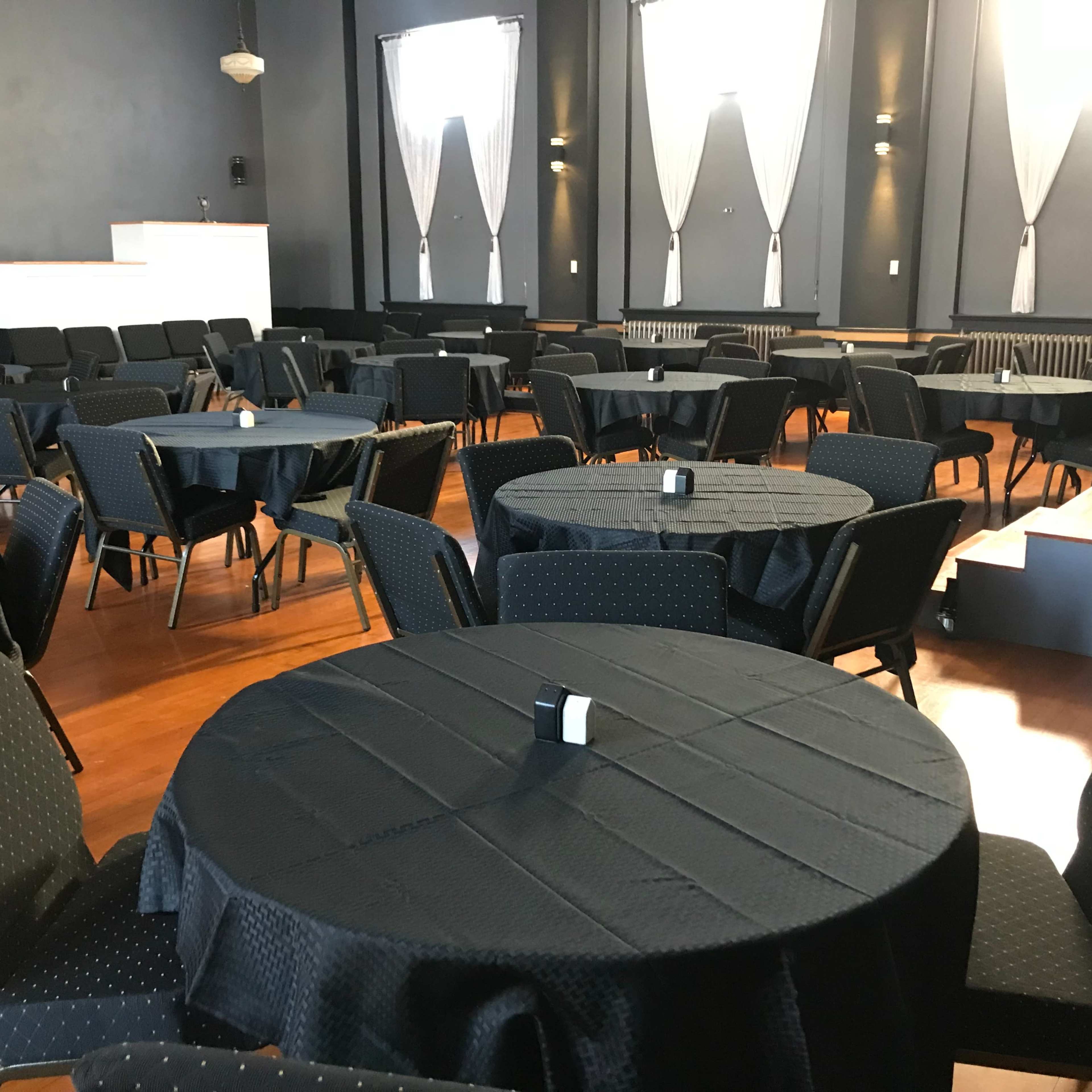 A spacious event hall set with round black tables surrounded by chairs, all arranged neatly on a polished wooden floor.
