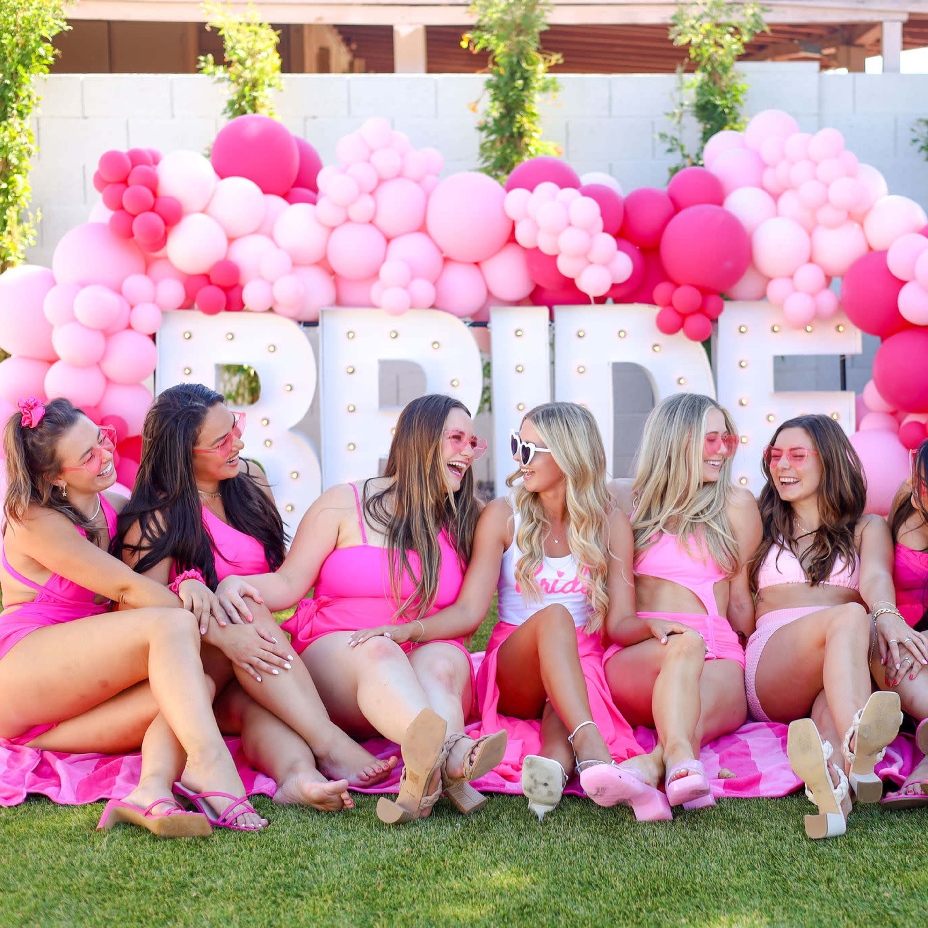 A group of women in pink swimsuits sit together on a blanket in front of a balloon arch that spells "BRIDE."