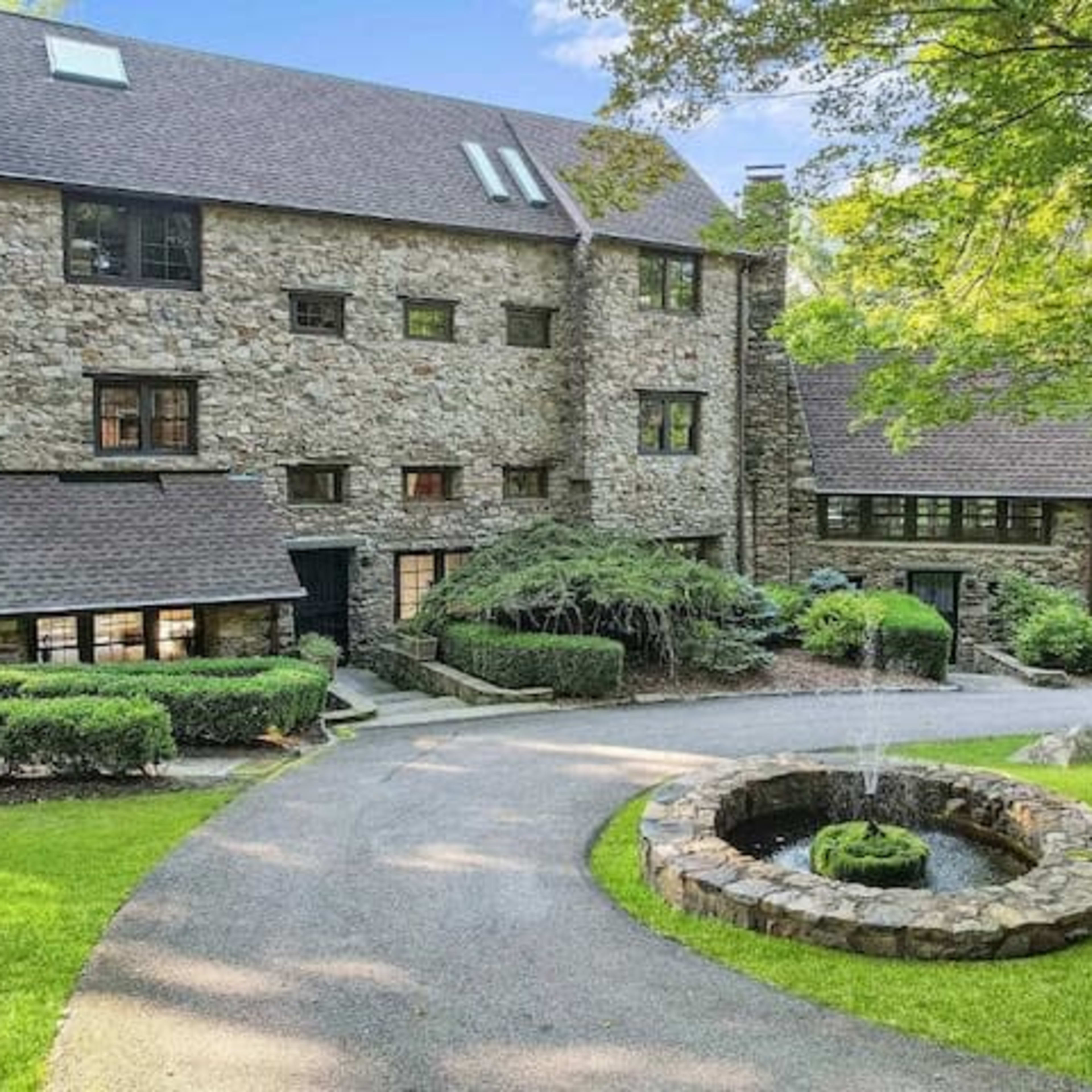 A multi-story stone house with a circular driveway and a fountain surrounded by a landscaped yard.