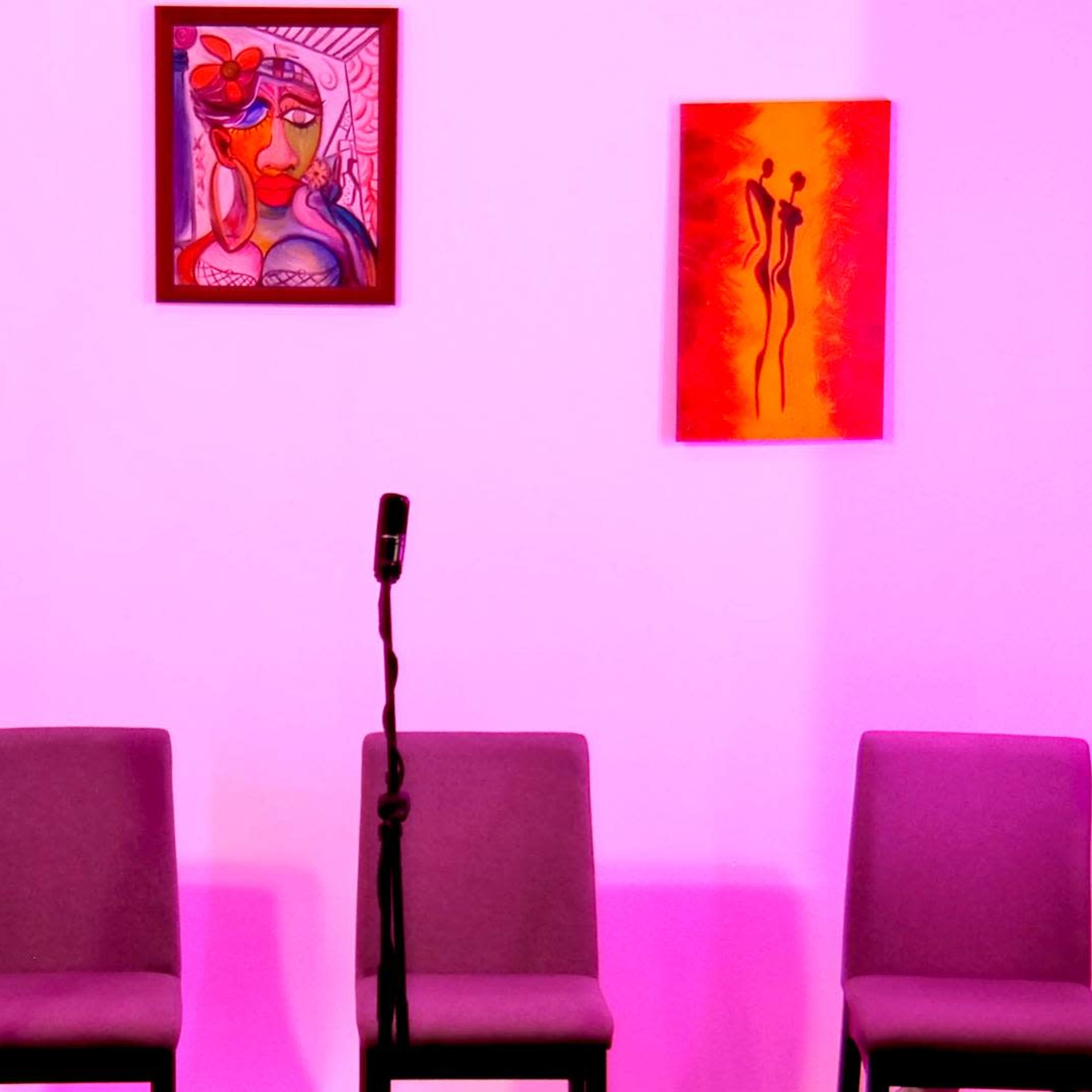 A stage setup with three empty chairs and two microphones, framed by colorful artwork on a pink-lit wall.