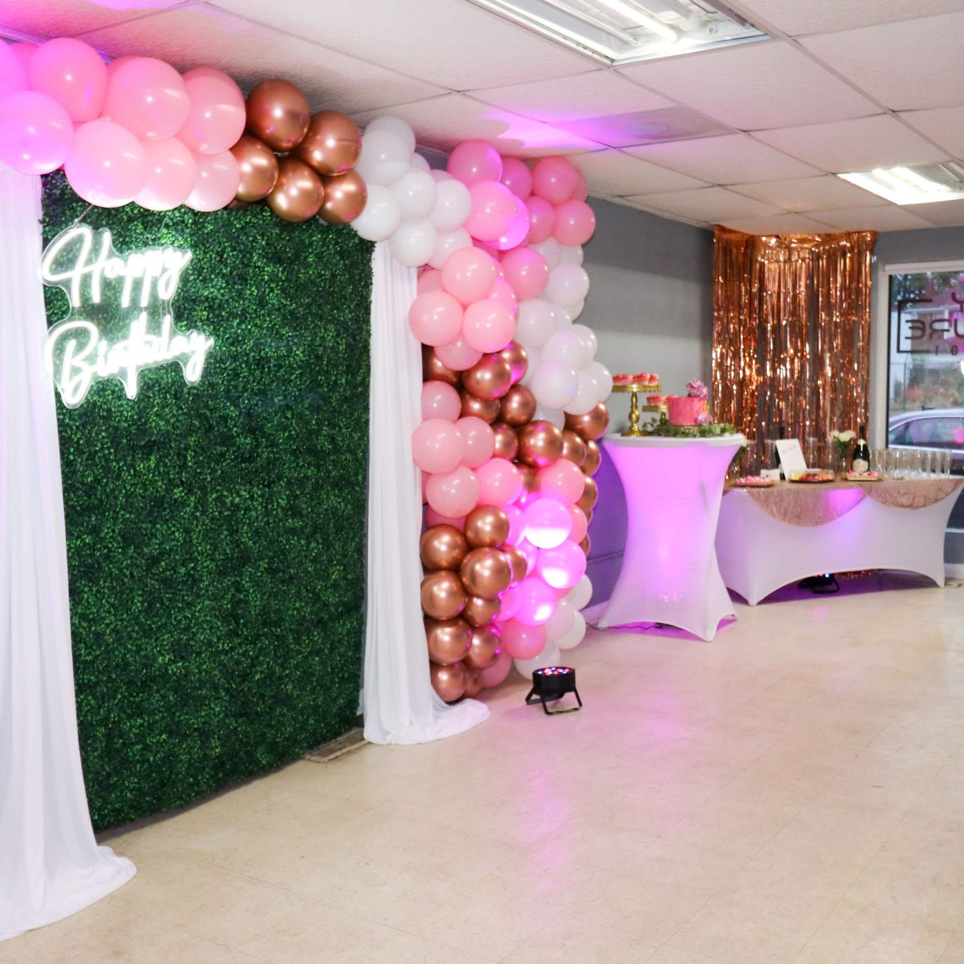 A decorated birthday celebration area featuring a green wall with "Happy Birthday" neon lettering, a balloon arch in pink and gold hues, and a refreshment table with metallic accents.