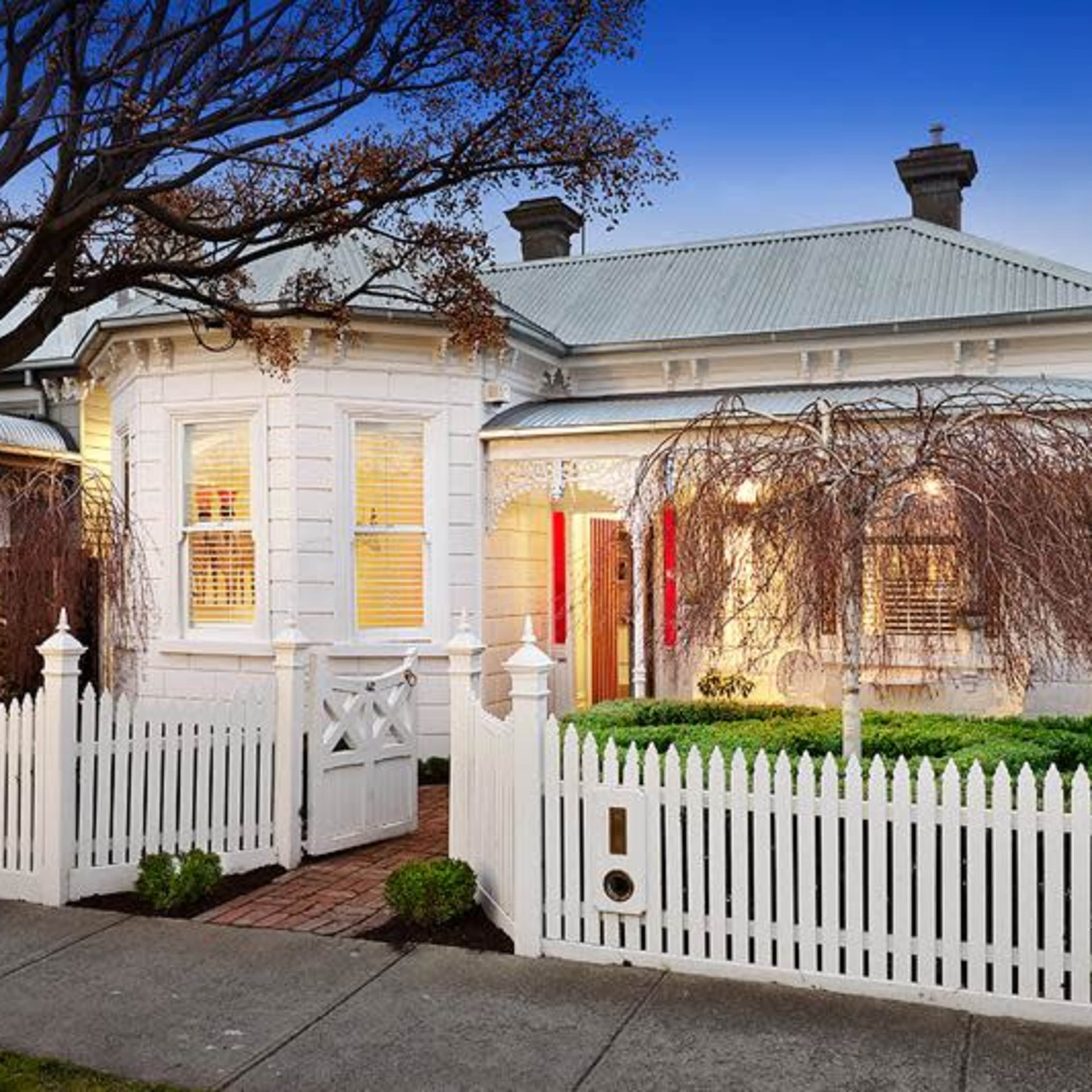 A white, single-story house with a curved facade, surrounded by a white picket fence and greenery, is shown at dusk.