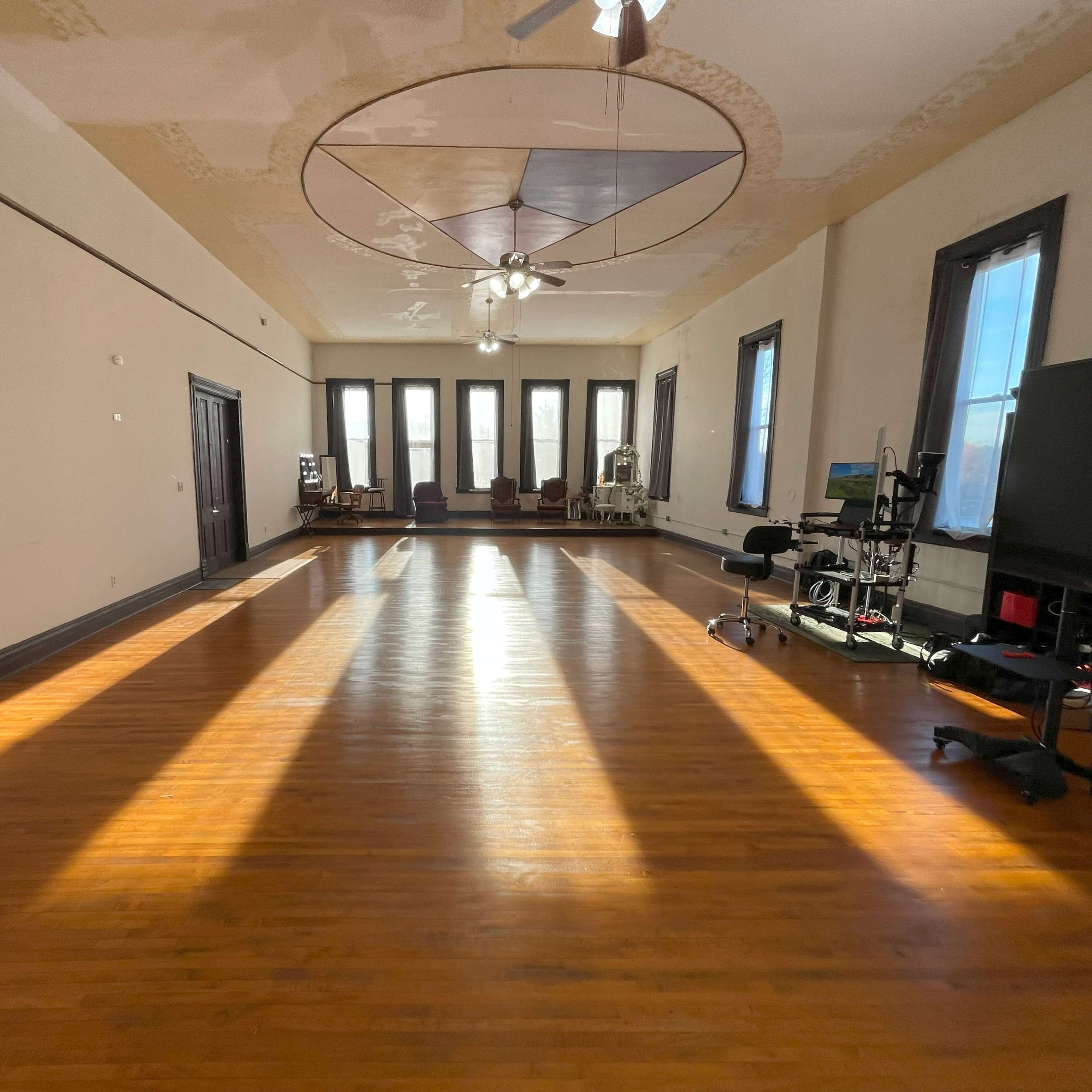 A spacious, well-lit room with wooden floors, large windows, and various pieces of furniture along the walls.