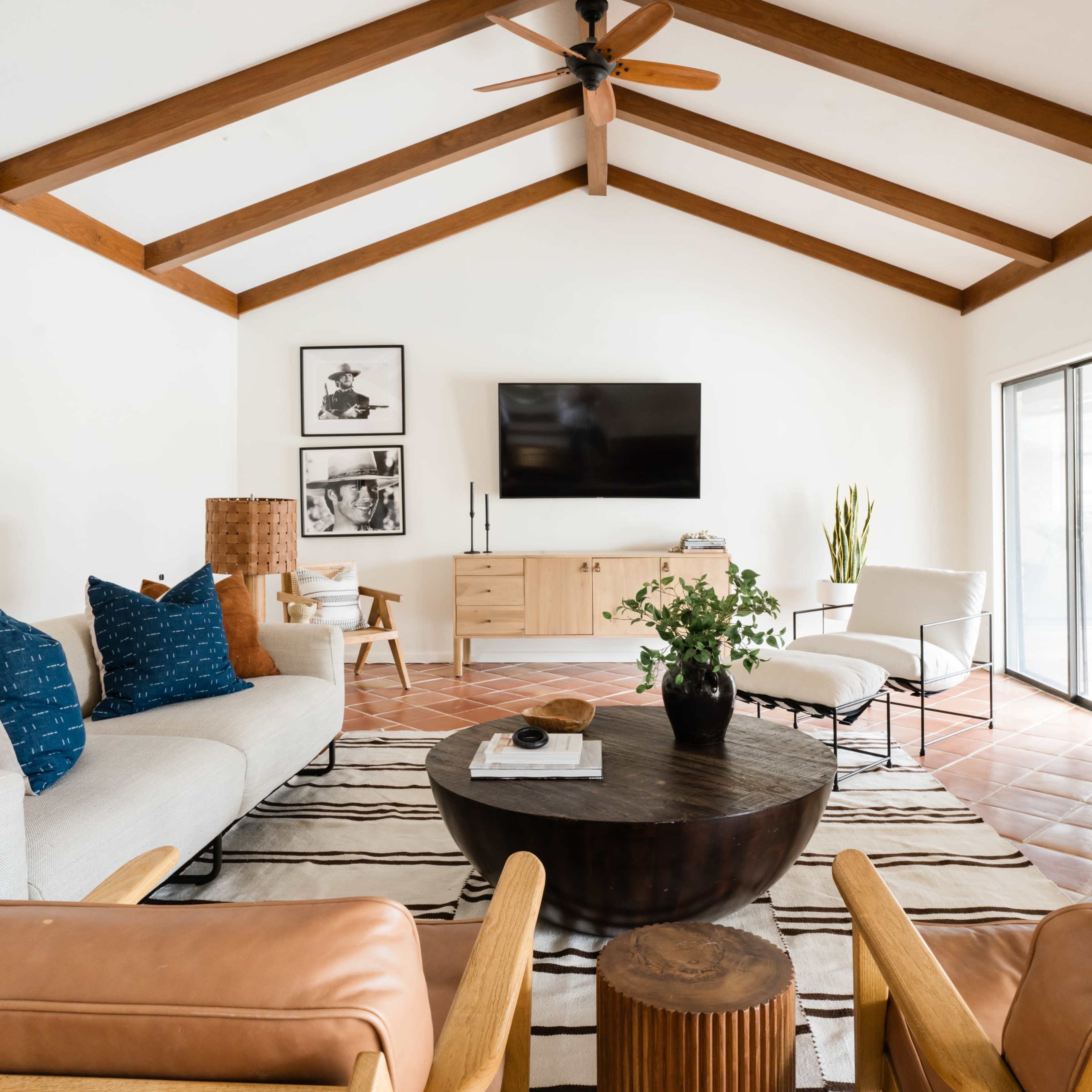 The living room features a sofa with blue and orange pillows, a round coffee table, and a wall-mounted television, all set beneath a vaulted wooden ceiling.