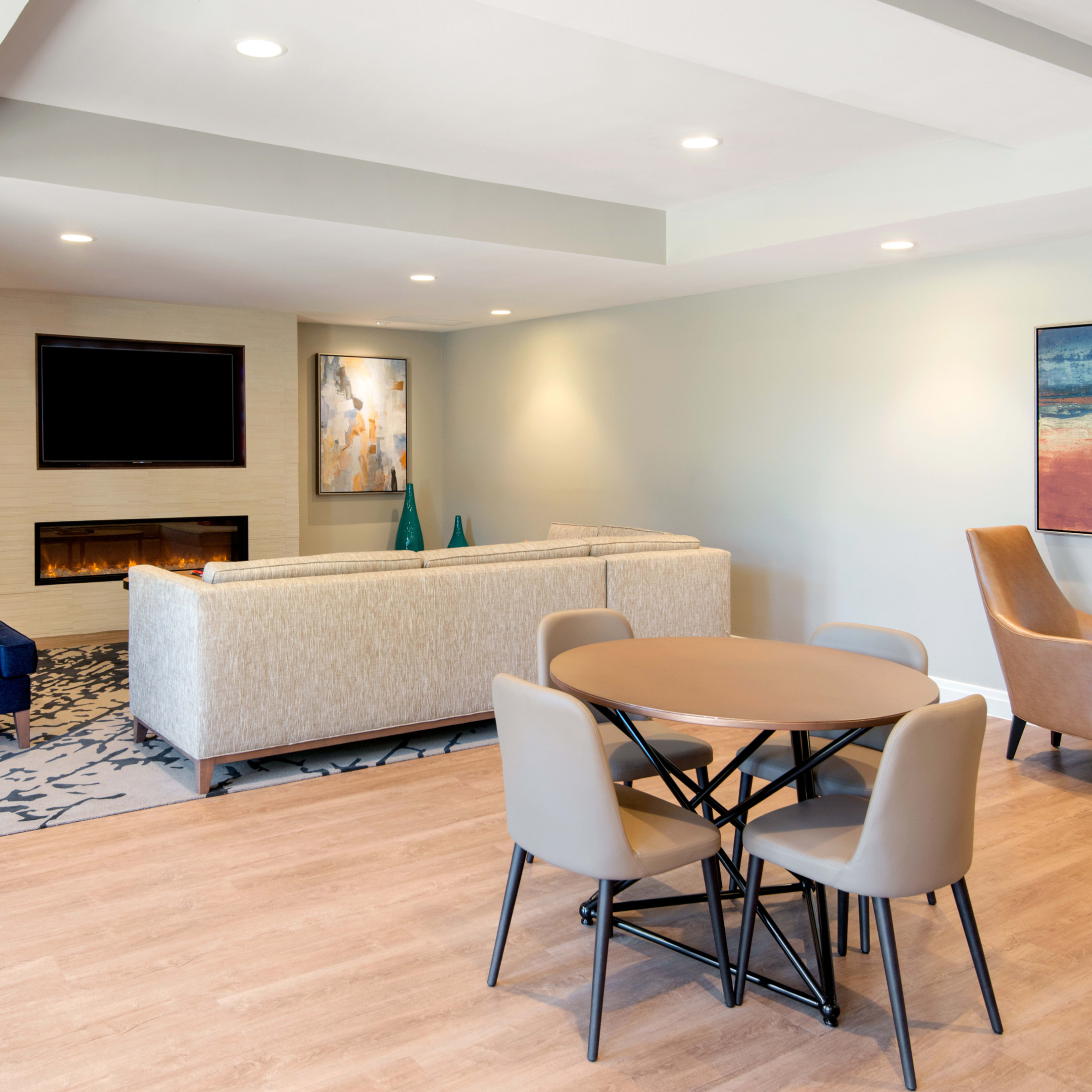 A modern living area features a sectional sofa, a round dining table with four chairs, and wall-mounted art on light-colored walls.