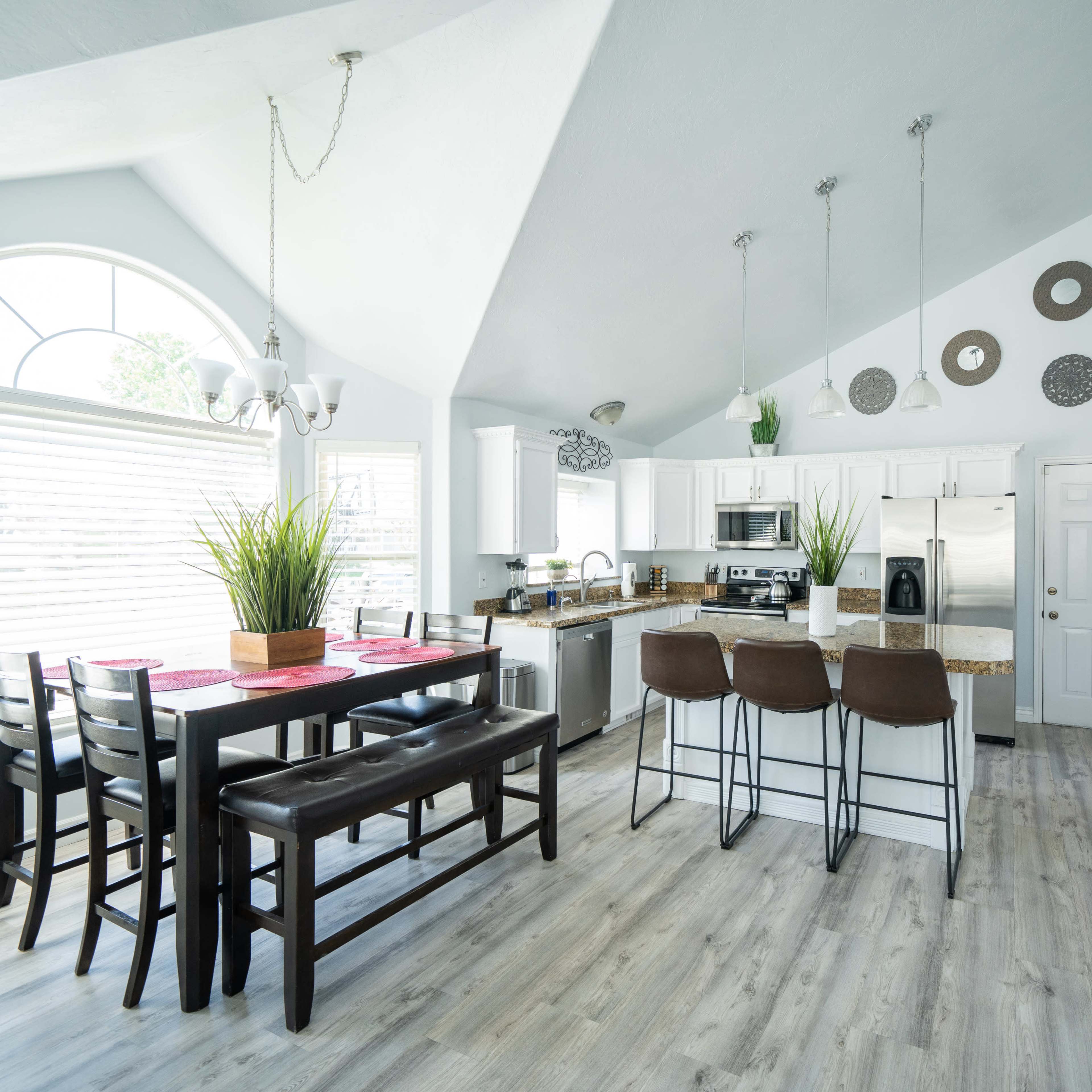 A modern kitchen with white cabinetry, a large dining table, and a bar area featuring three brown stools.