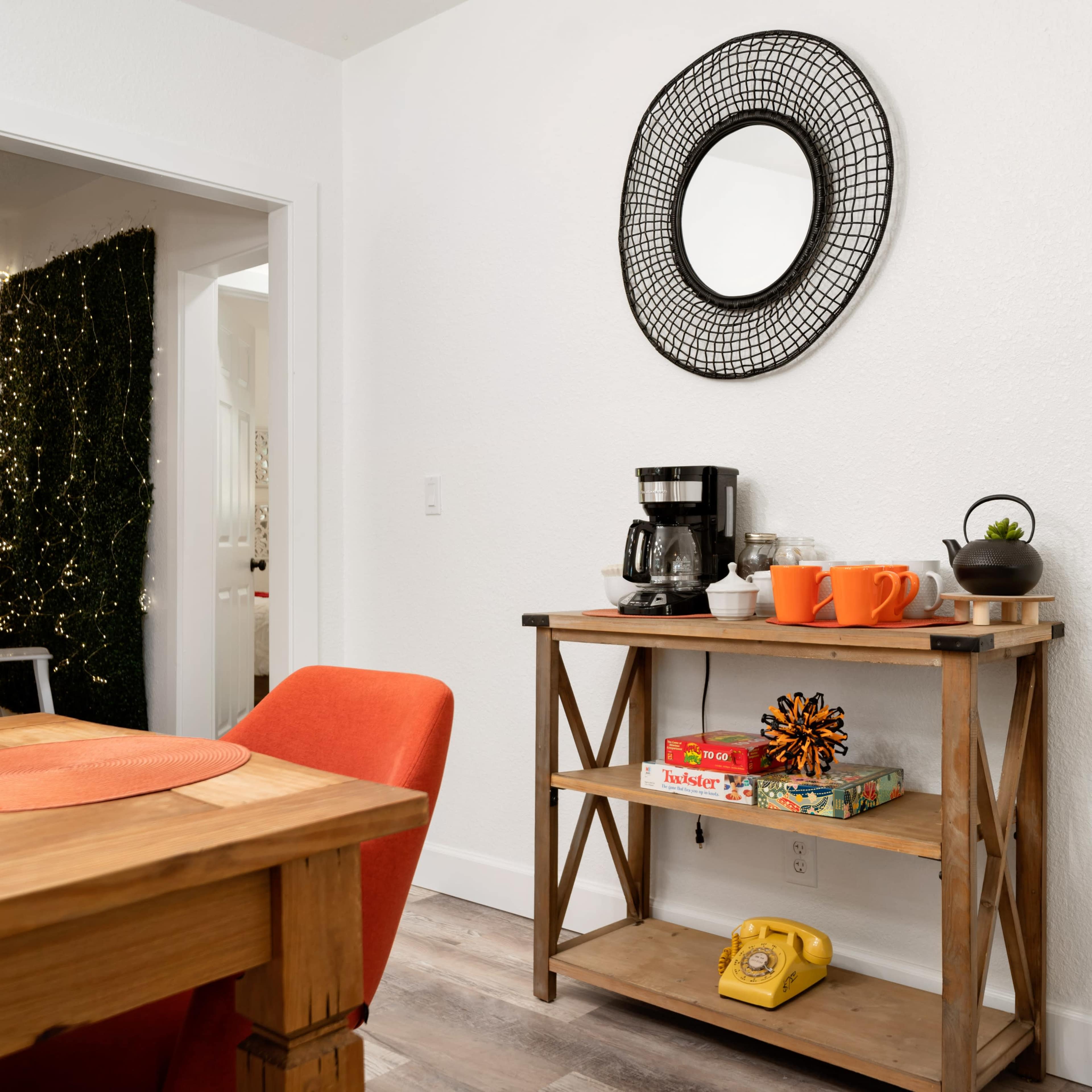A wooden table with an orange chair is positioned next to a shelf displaying a coffee maker, orange mugs, a small plant, and a vintage yellow telephone, with a round mirror mounted on the wall.