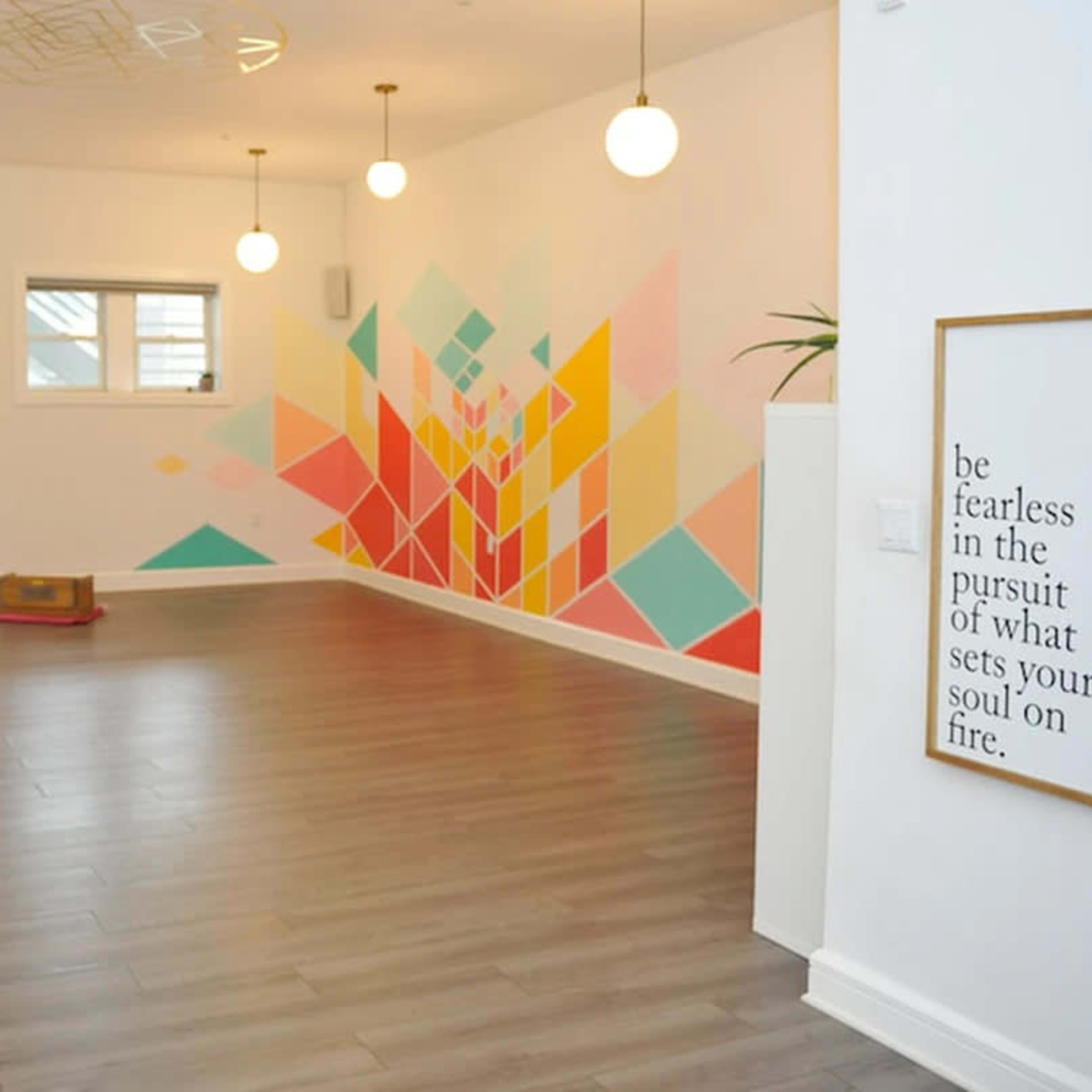 The room features a light wooden floor, a colorful geometric mural on the wall, and pendant lights hanging from the ceiling.