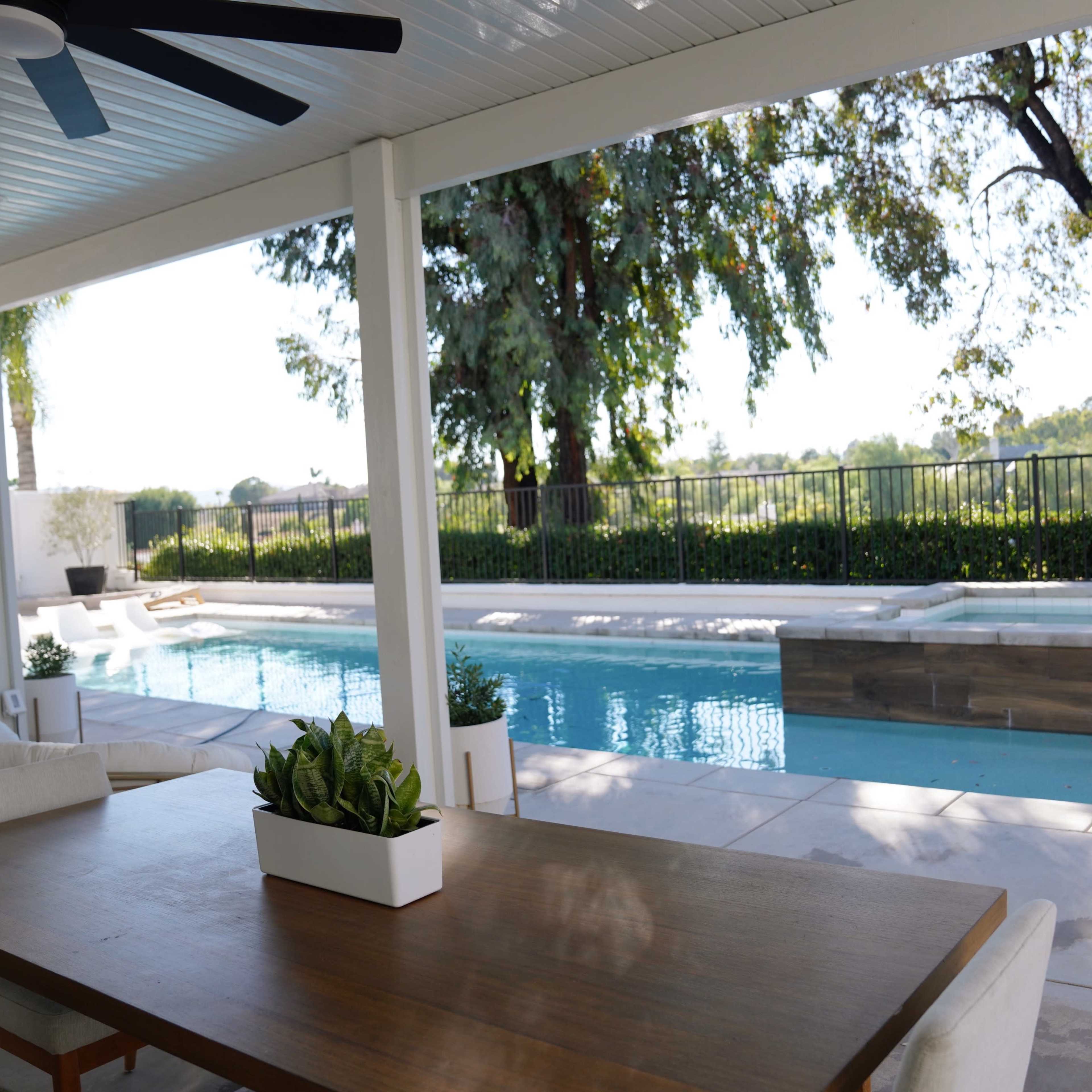 A spacious patio features a dining table overlooking a swimming pool surrounded by greenery and a fence.