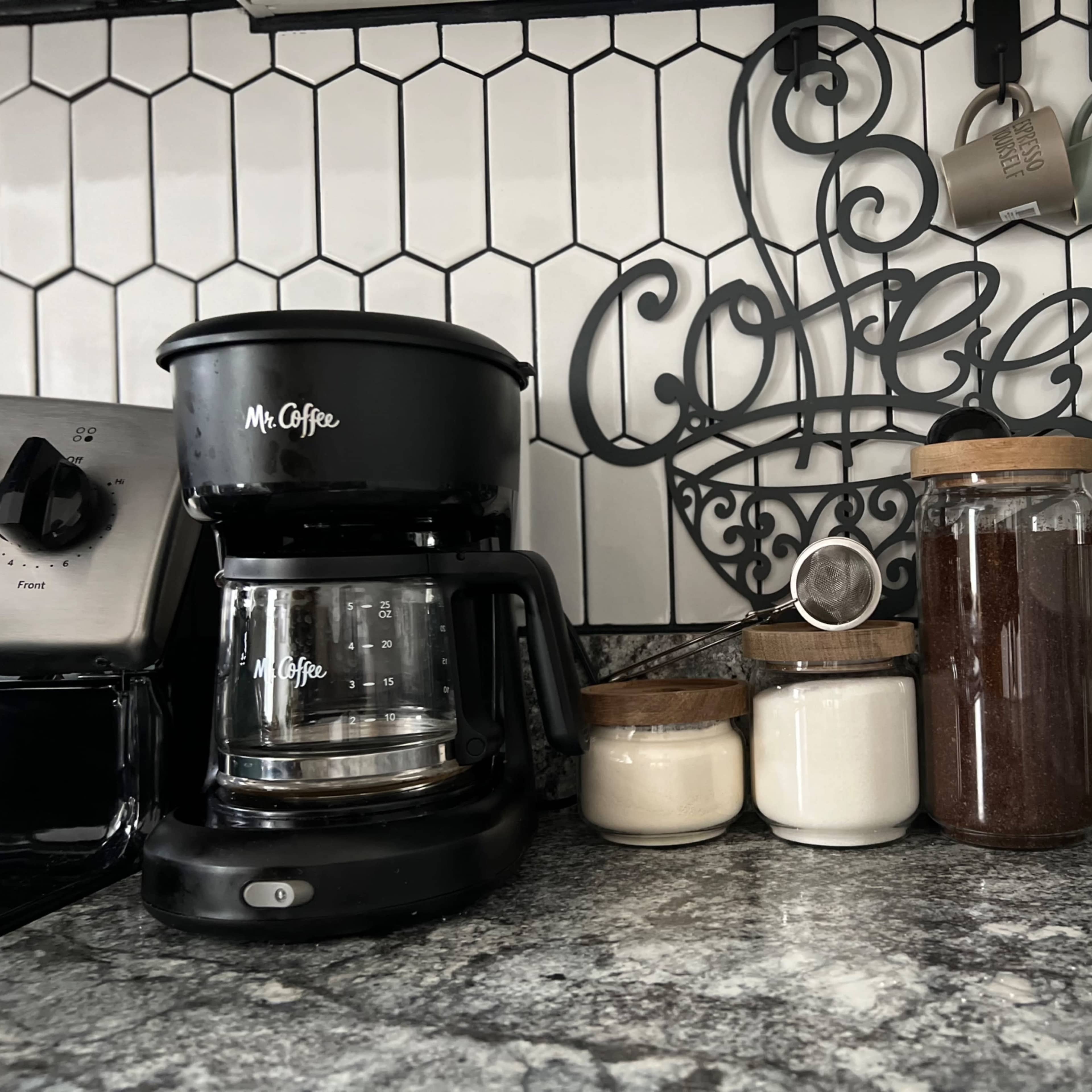 A coffee station features a Mr. Coffee maker, jars of sugar and coffee, and a French press on a granite countertop, with a decorative coffee sign above.