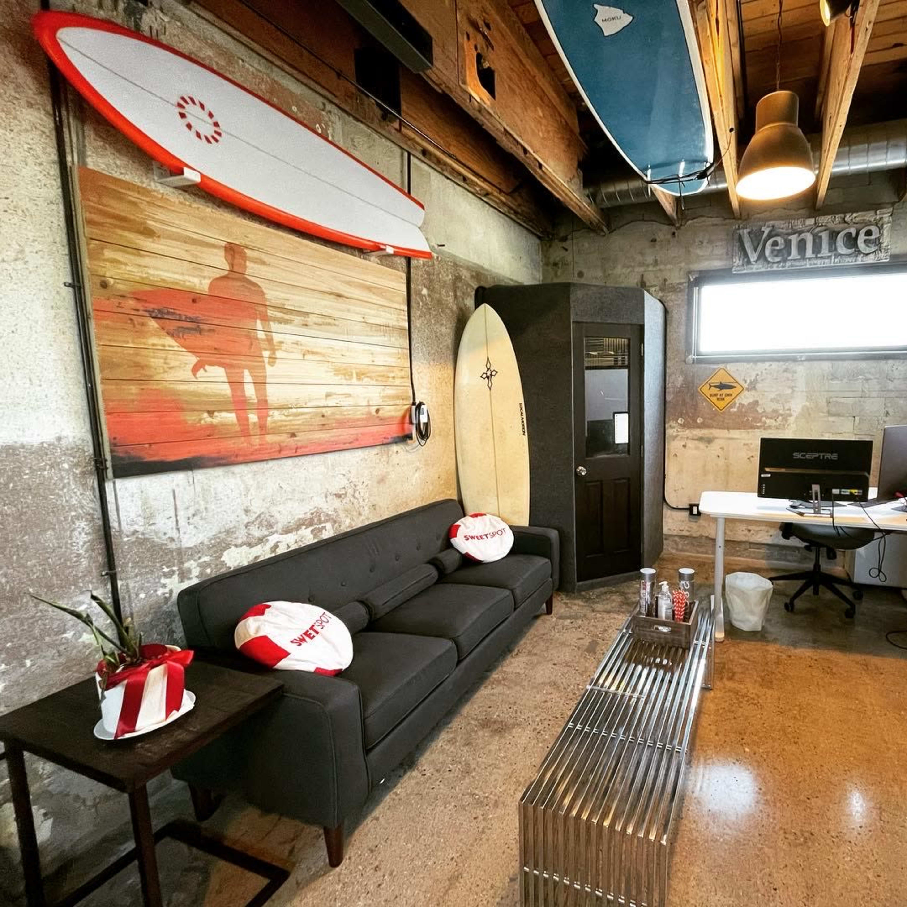 A contemporary workspace with a gray sofa, a surfboard mounted on the wall, and a desk setup with a computer and television in a minimalist room with exposed concrete walls.