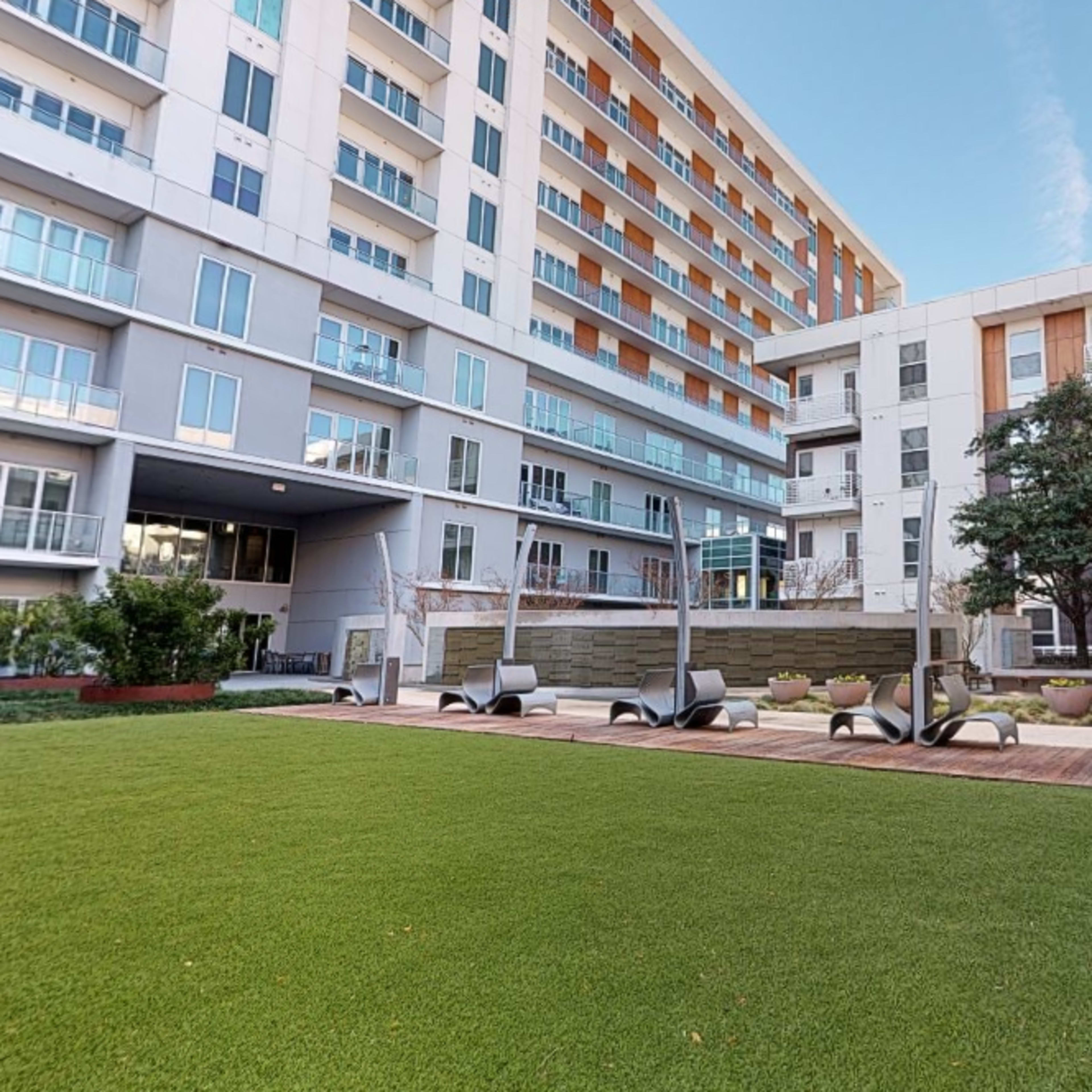 A landscaped courtyard area featuring artificial grass, seating arrangements, and several modern apartment buildings in the background.
