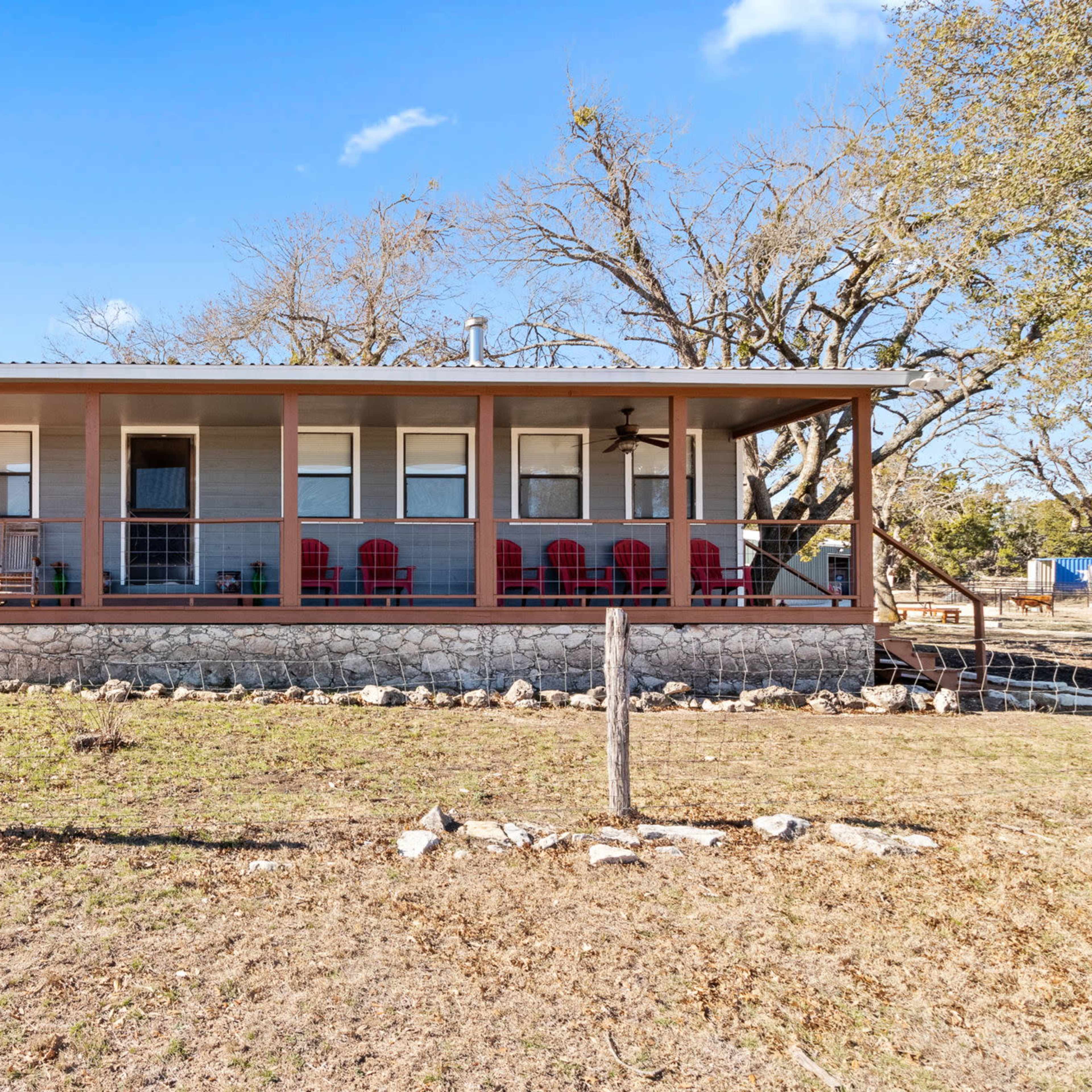 A single-story house with a covered porch, featuring several red chairs and surrounded by a grassy area and sparse trees.